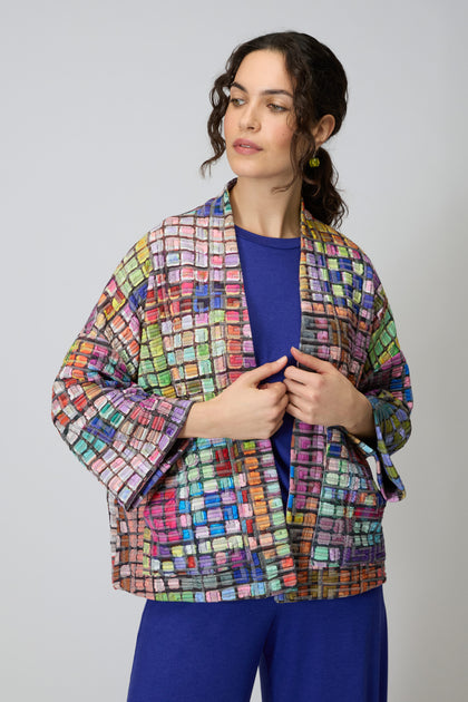 A woman models the Coloured Glass Tiles Jersey Jacket, featuring a vibrant mosaic pattern, over a blue top and blue pants against a plain light background.
