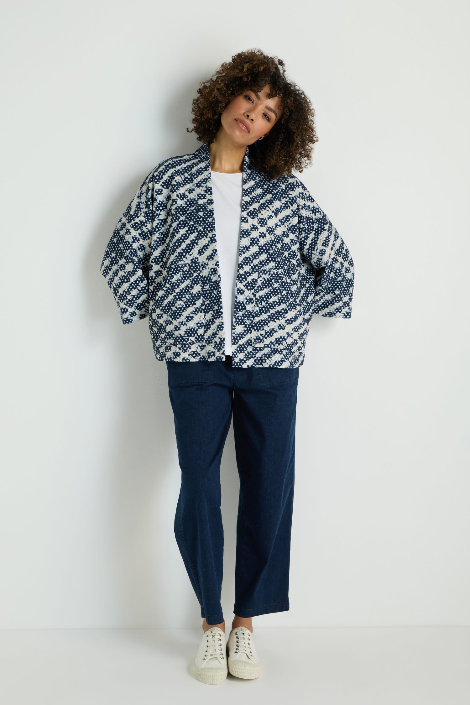 A person stands against a plain white background wearing a white T-shirt, navy blue pants, white sneakers, and the Diamond Tie Dye Jersey Jacket.