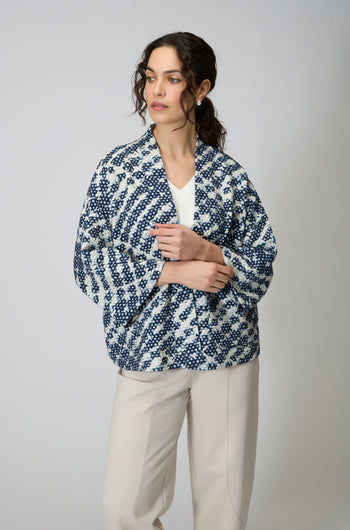 A woman stands against a plain background wearing the Diamond Tie Dye Jersey Jacket over a white top and beige pants.