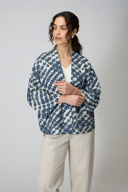 A woman stands against a plain background wearing the Diamond Tie Dye Jersey Jacket over a white top and beige pants.