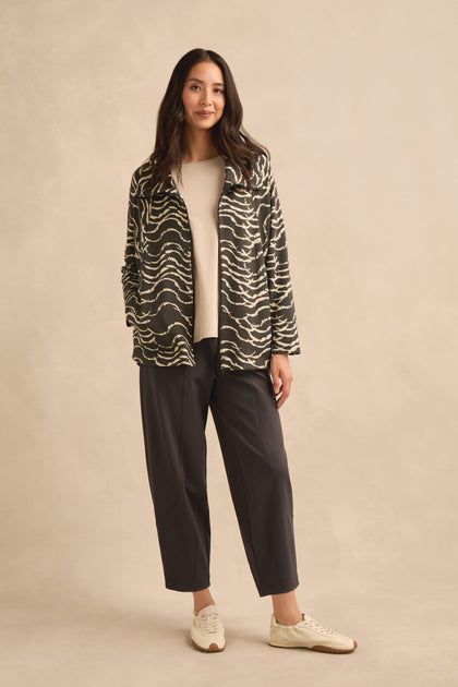 A woman stands against a beige background wearing the Dancing Waves Zip Boxy Jacket—a black and white wave motif jersey piece—paired with a cream top, dark cropped pants, and white sneakers.