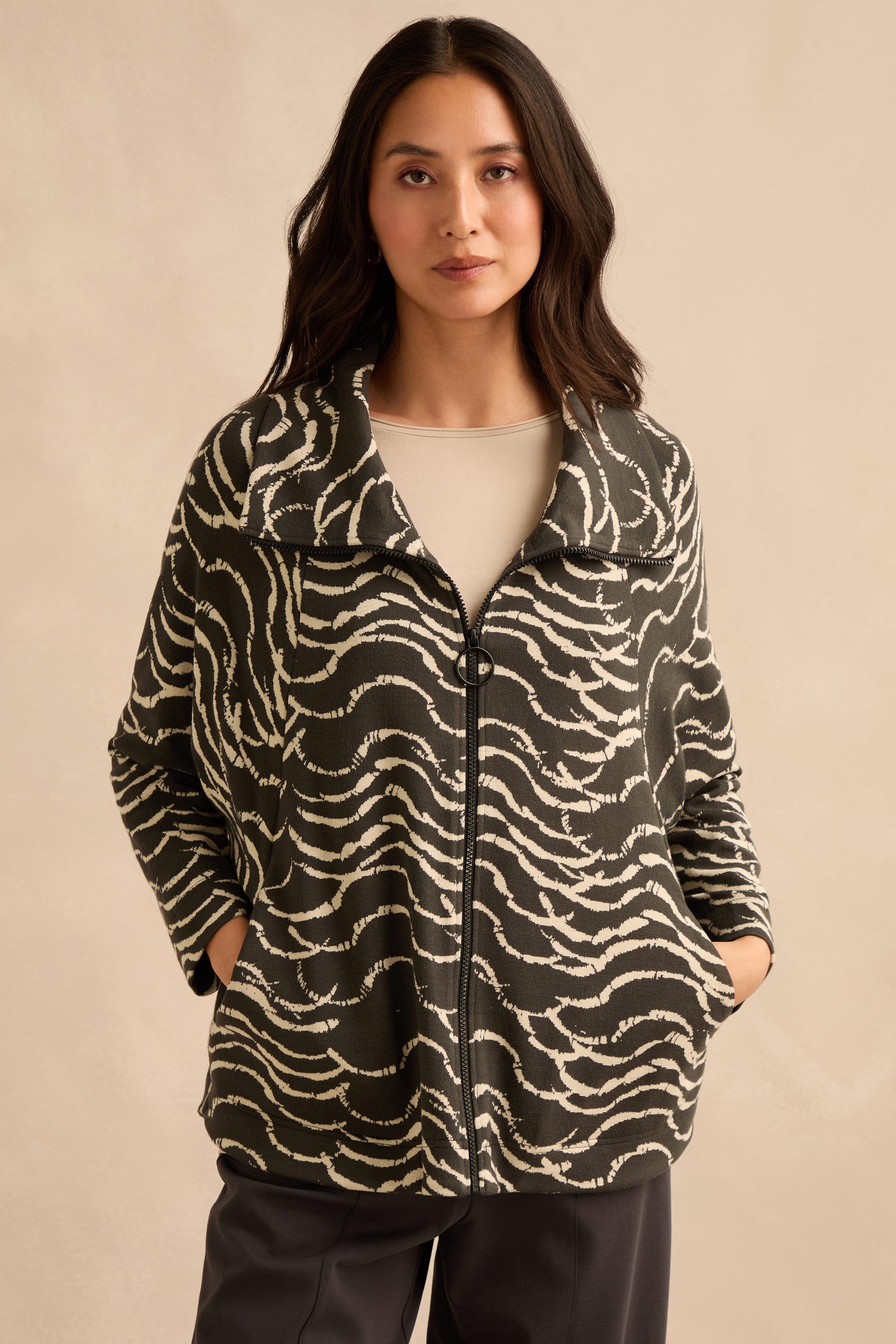 A woman stands with her hands in the pockets of a Dancing Waves Zip Boxy Jacket—featuring a black and white wave motif—paired with a light beige top and dark pants, against a plain beige background.
