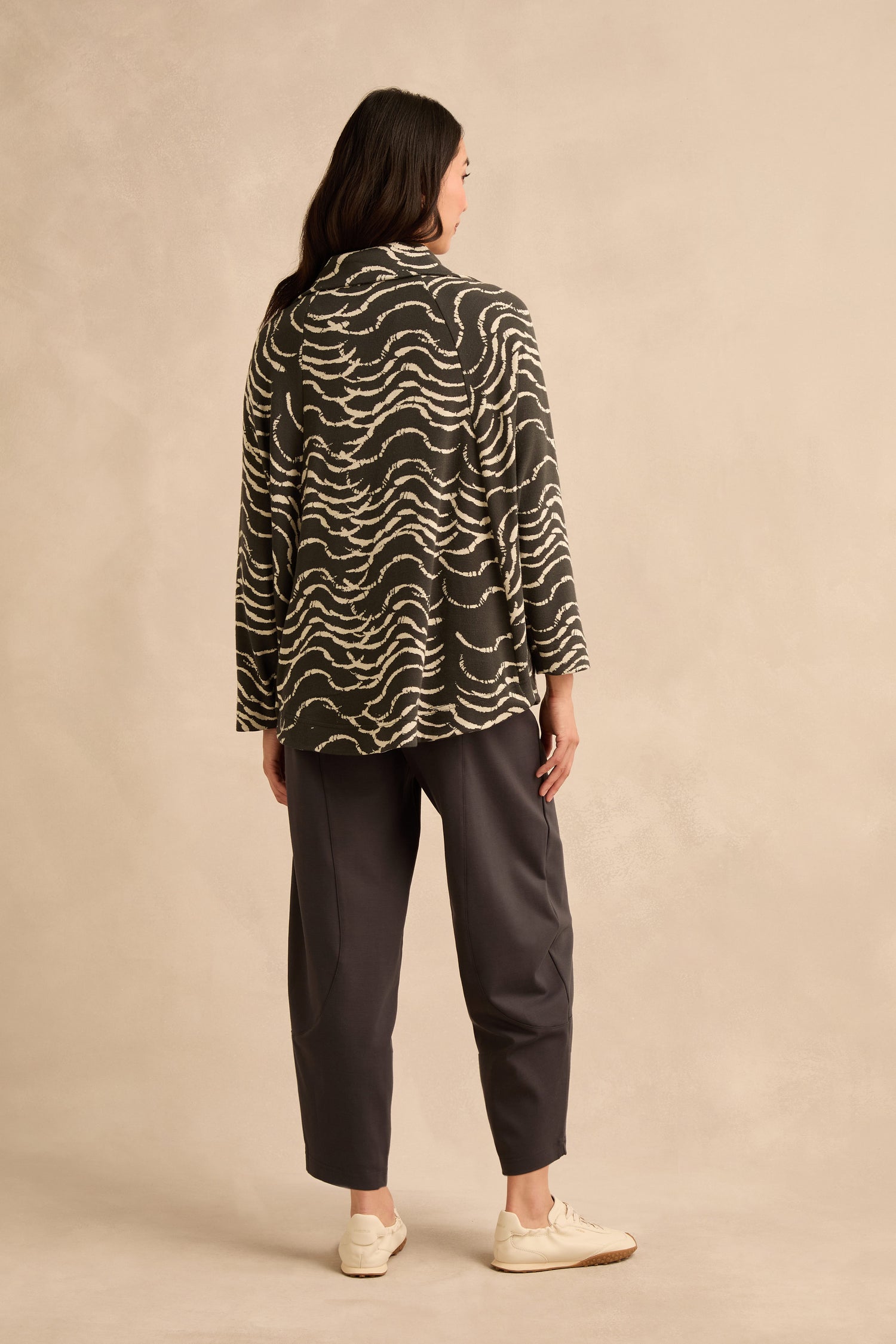 A person stands with their back to the camera, wearing the Dancing Waves Zip Boxy Jacket—characterized by its long sleeves and wave motif—paired with dark loose pants and white sneakers, set against a neutral background.
