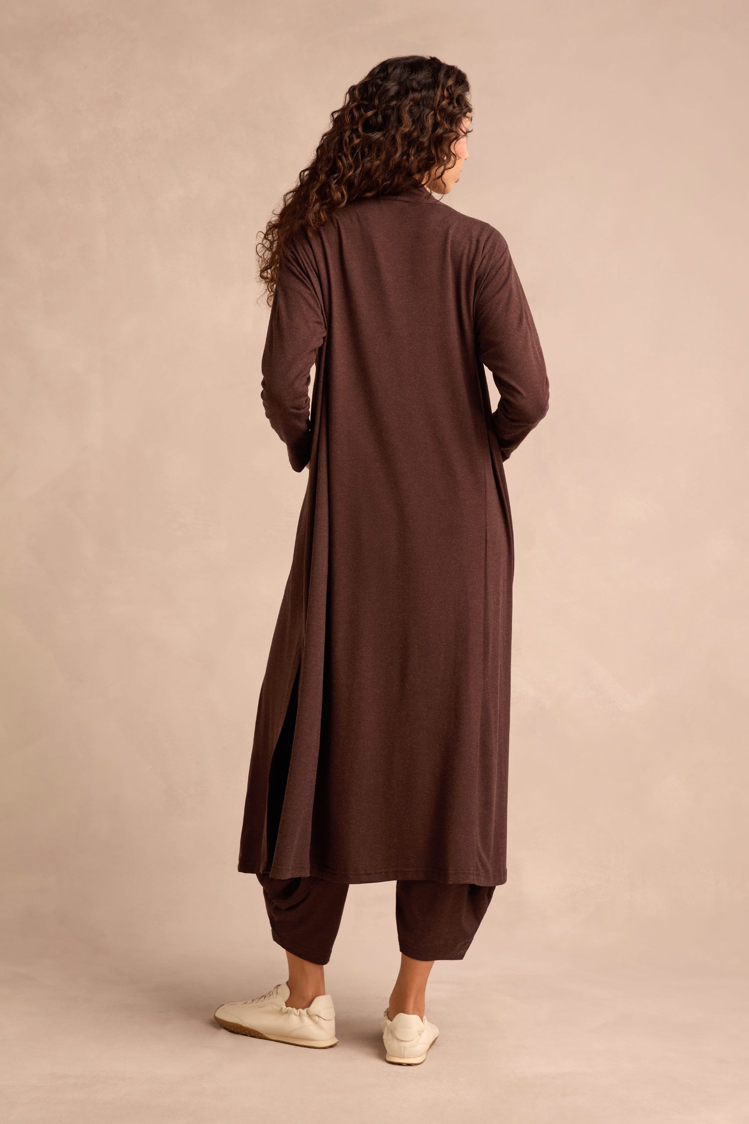 A person stands with their back to the camera, wearing the Soft Viscose Jersey Long Line Cardi in rich brown, paired with matching brown pants and white sneakers against a neutral backdrop.