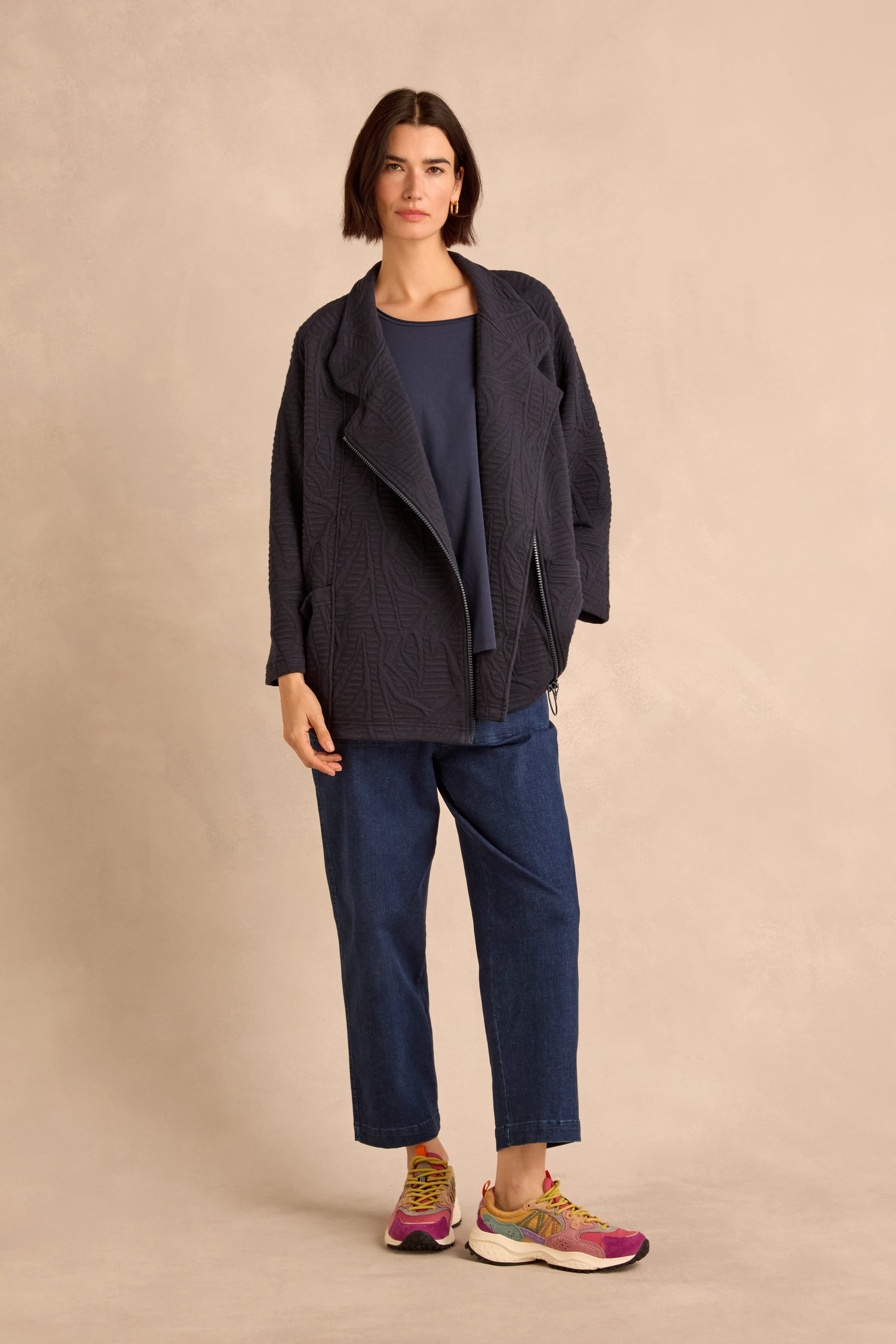 Woman standing against a beige background, wearing the Quilted Zip Through Jacket in navy cotton-rich fabric made in England, paired with a blue top, loose blue jeans, and colorful sneakers.