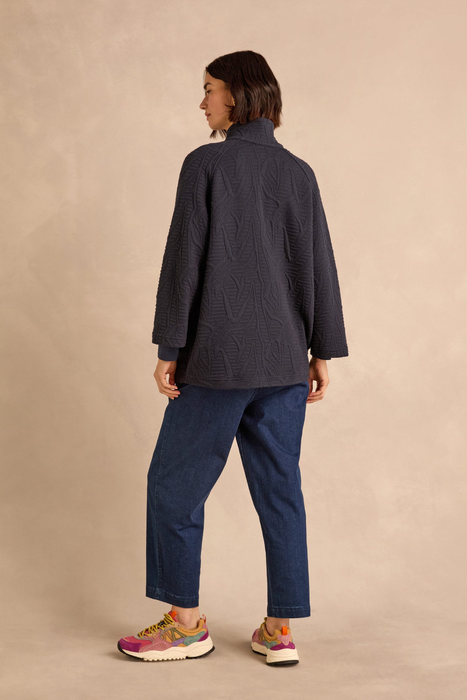 A person stands facing away, wearing the Quilted Zip Through Jacket with a textured cotton-rich finish, blue jeans, and colorful sneakers against a beige background.