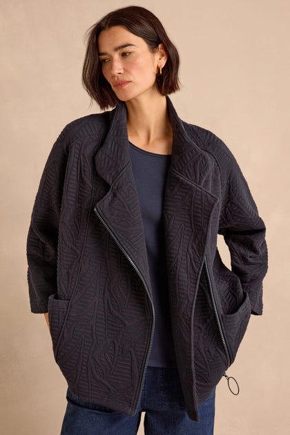 Woman wearing the Quilted Zip Through Jacket, featuring a textured, cotton-rich design with leaf patterns, over a navy top and jeans, standing against a plain beige background.