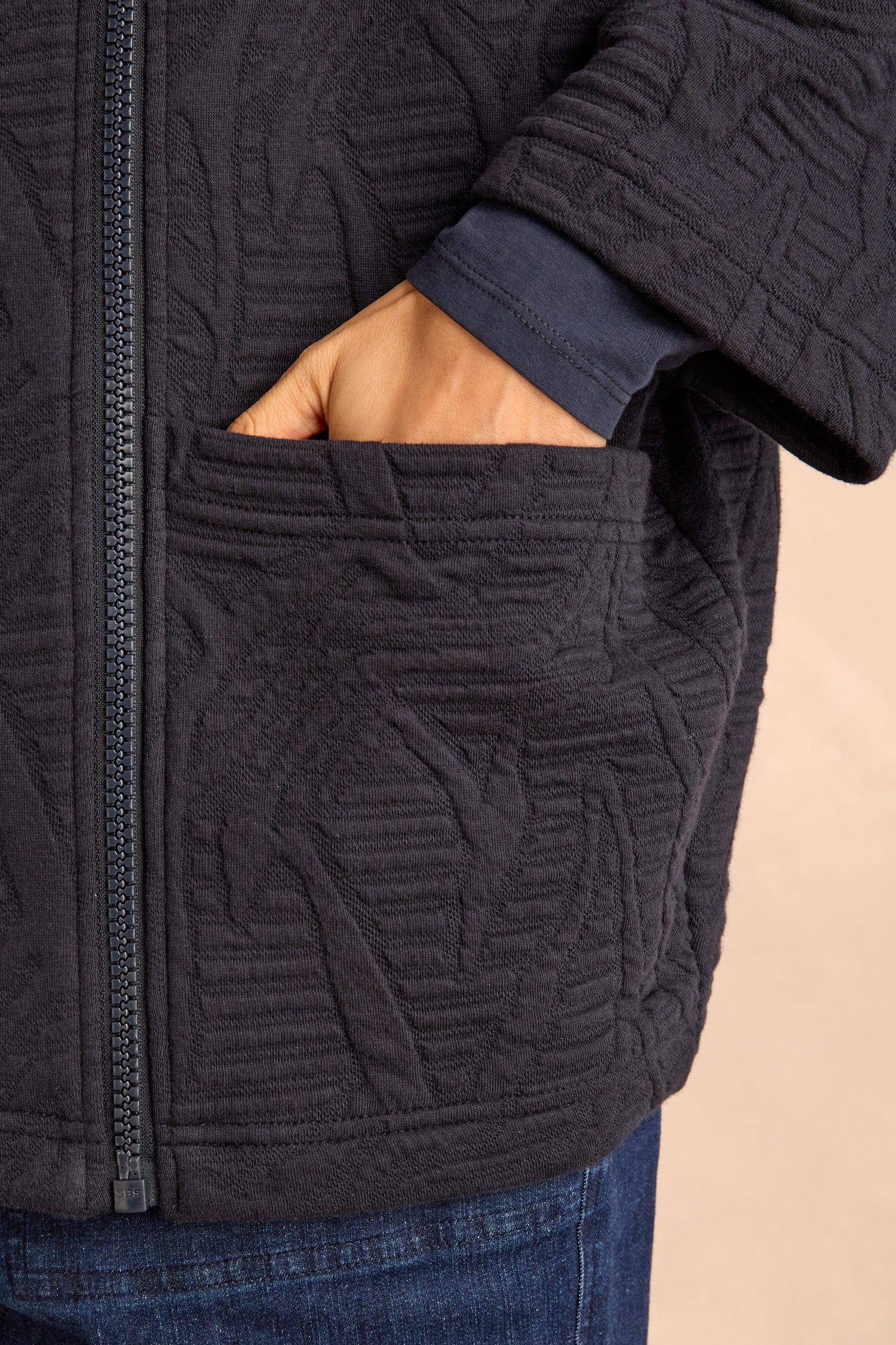 A person wearing a dark Quilted Zip Through Jacket and blue jeans has their right hand in a front pocket.