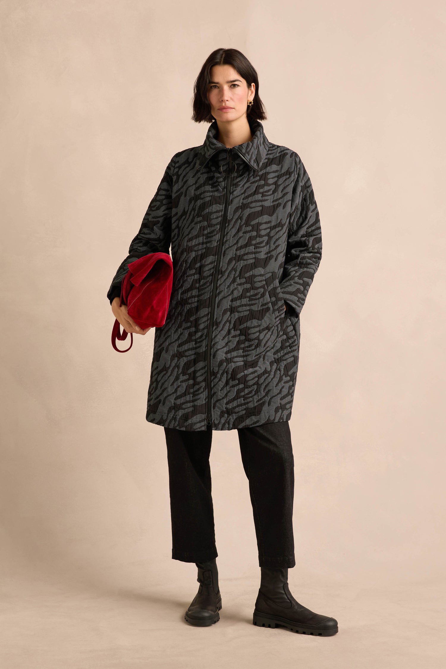 Wearing the Quilted Coat, a person stands against a neutral background, showcasing its oversized silhouette and knee-length design. The look is completed with black pants, boots, and a red bag.