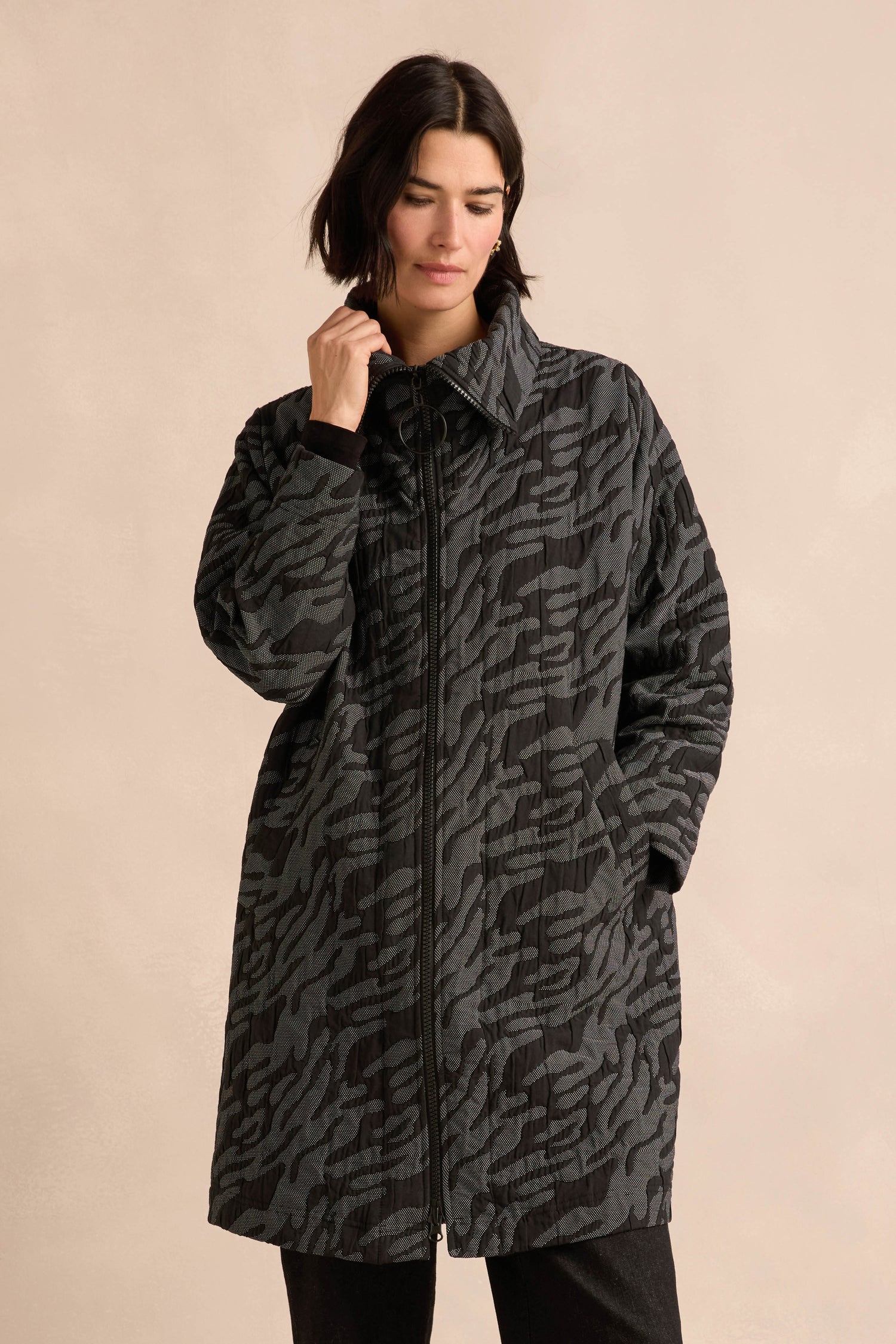 A person wearing the Quilted Coat, featuring a long, dark, patterned design with an oversized silhouette and hands in pockets, stands against a plain beige background.