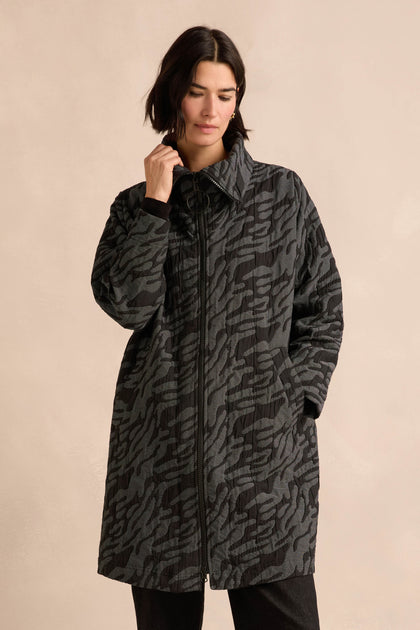 A person wearing the Quilted Coat, featuring a long, dark, patterned design with an oversized silhouette and hands in pockets, stands against a plain beige background.