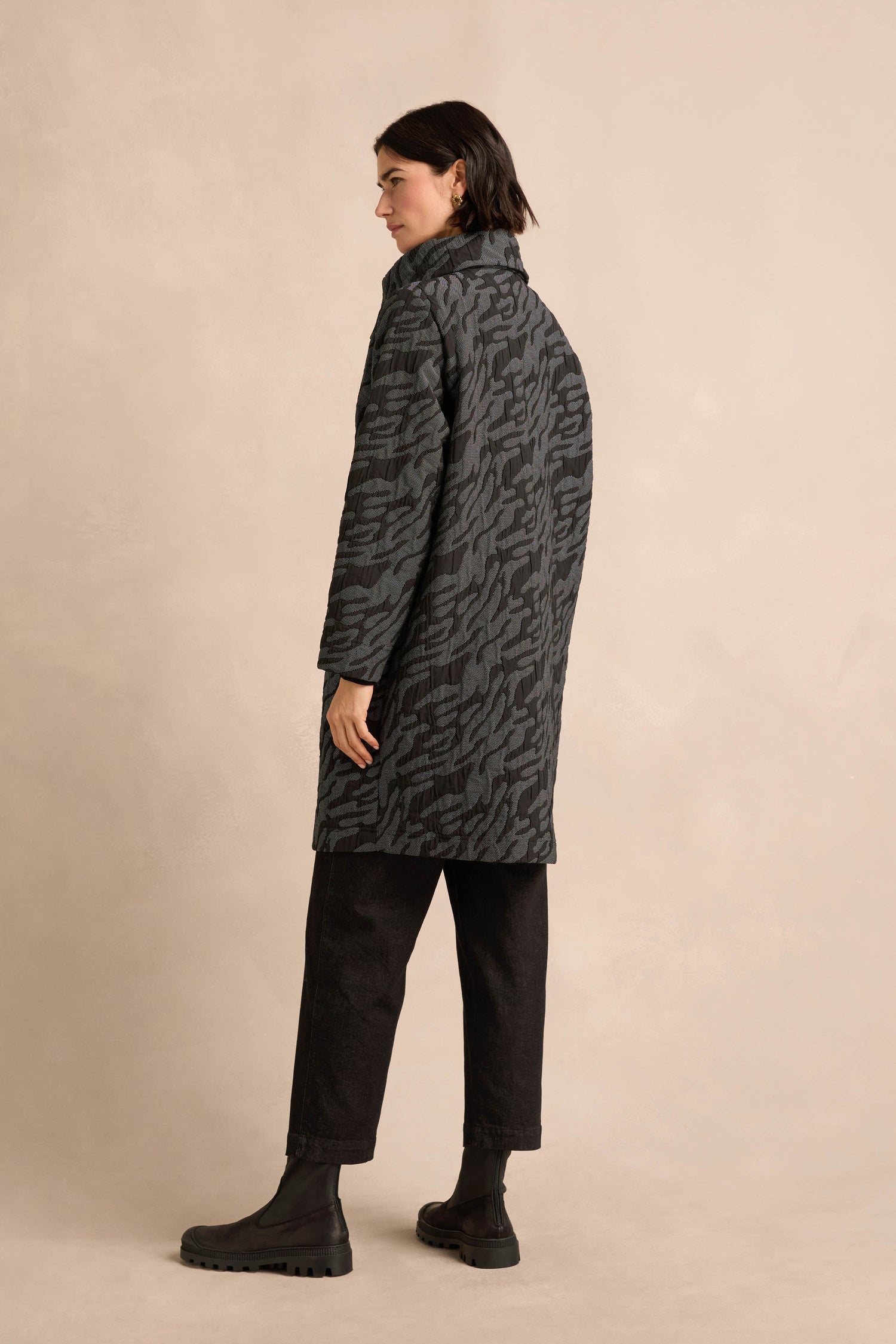 A person stands facing away, wearing the Quilted Coat, which features a dark patterned quilted design and an oversized silhouette, paired with black pants and black boots against a light beige background.