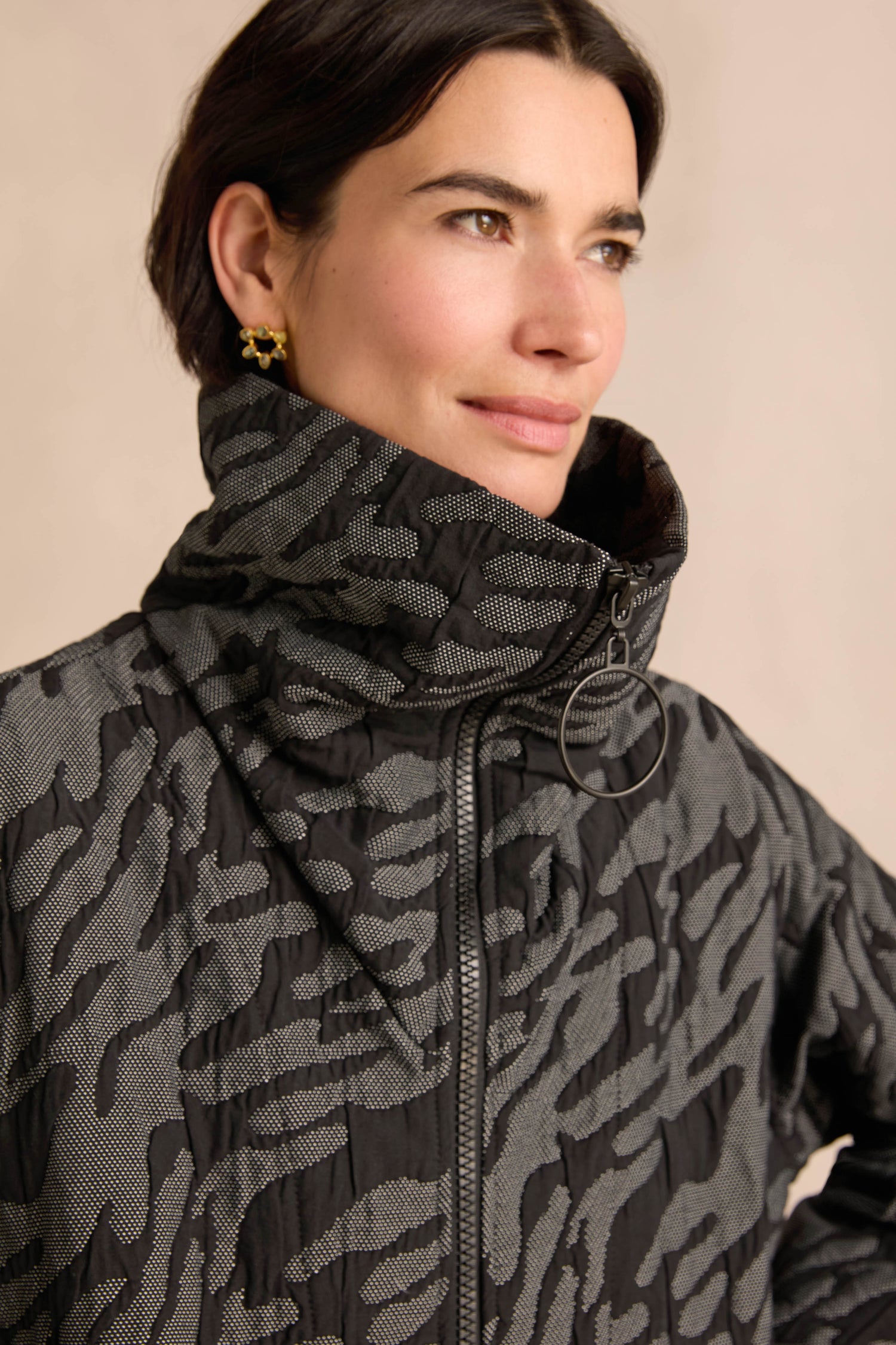 A woman with short dark hair stands against a beige background wearing the Quilted Coat, featuring a high collar, black and gray patterned quilting, an oversized silhouette, and a distinctive hoop zipper detail. She completes her look with gold star earrings.