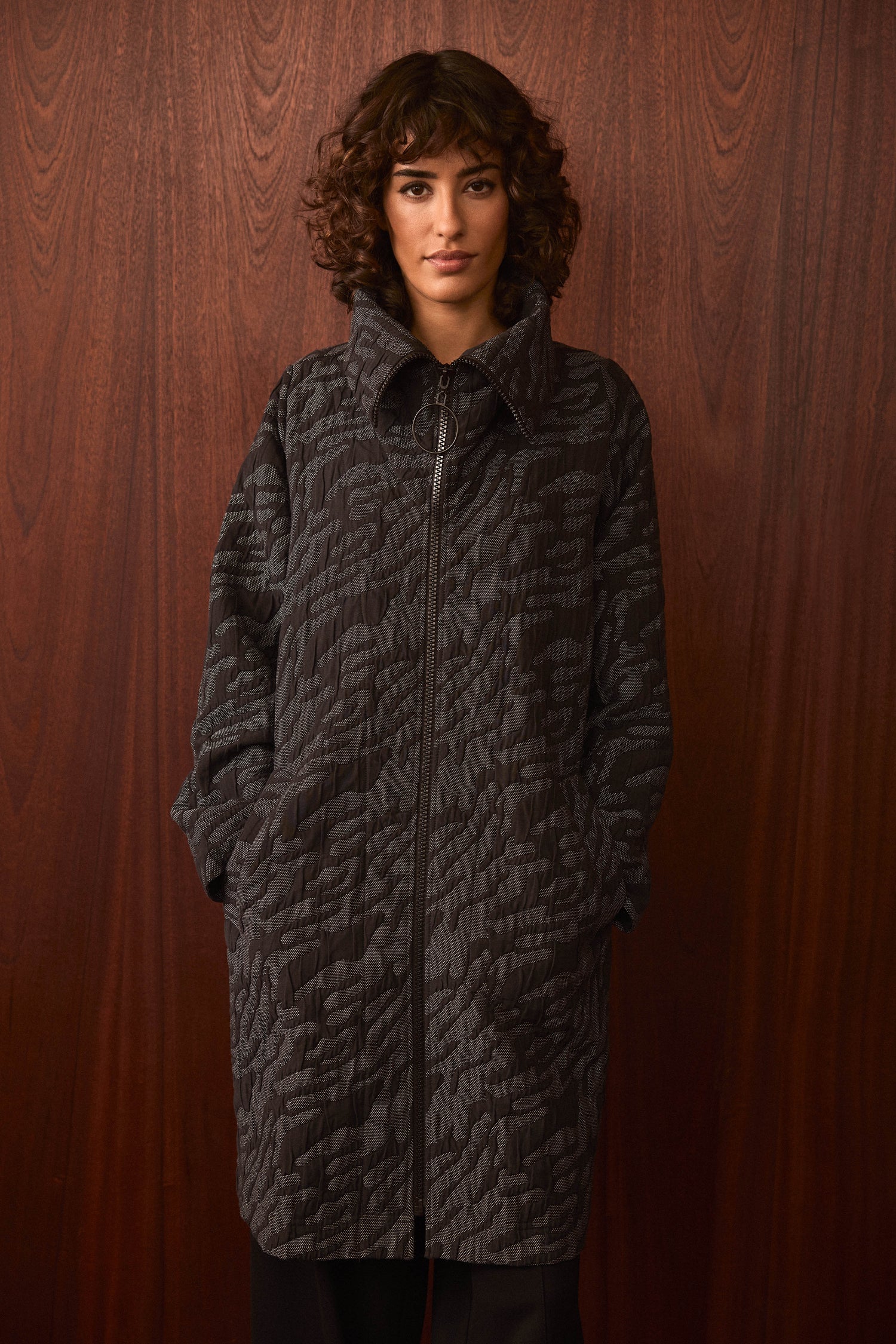 A person with curly hair wears the Quilted Coat, style SPJ7062-QDC—an oversized, textured dark coat—with hands in the pockets, standing against a wooden wall background.