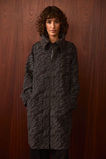 A person with curly hair wears the Quilted Coat, style SPJ7062-QDC—an oversized, textured dark coat—with hands in the pockets, standing against a wooden wall background.