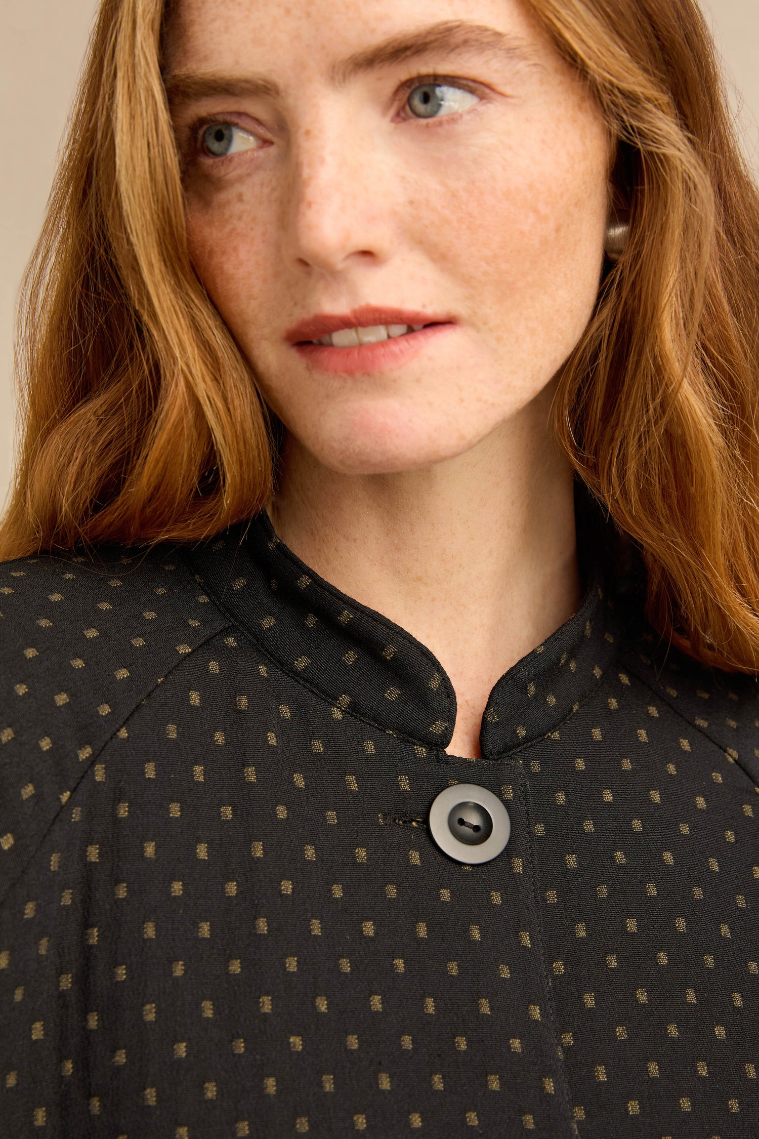 A woman with red hair and freckles wears the Square Jacquard Jacket, a workwear-inspired black blouse featuring a gold dot pattern and a mandarin collar fastened with a grey button.