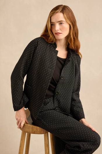 A woman sits on a wooden stool wearing a workwear-inspired Square Jacquard Jacket with matching trousers and a black top underneath.