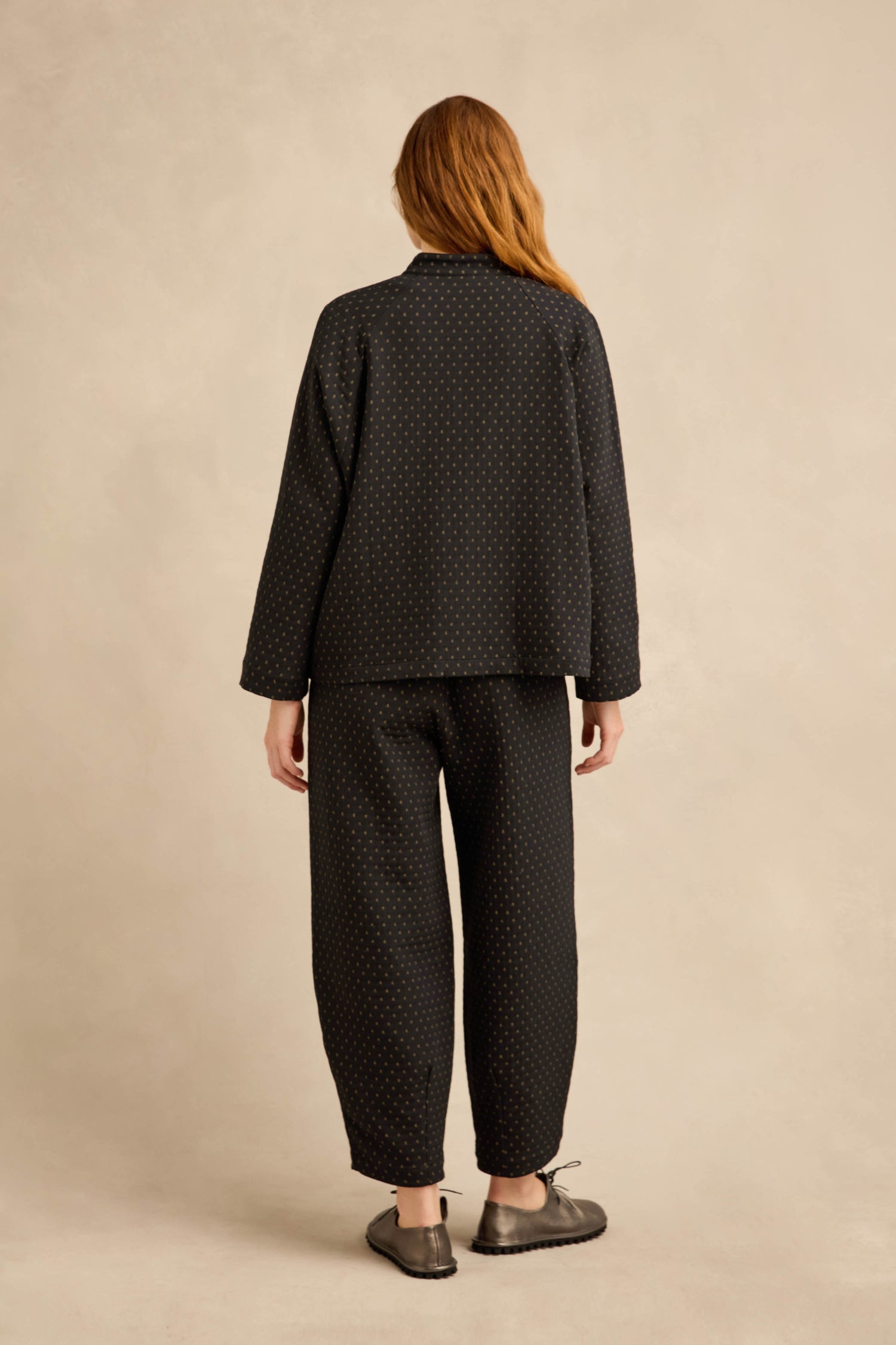 A person stands with their back to the camera, wearing a black long-sleeve shirt and dotted pants layered under the Square Jacquard Jacket for a modern, workwear-inspired look, paired with gray slippers against a plain beige background.