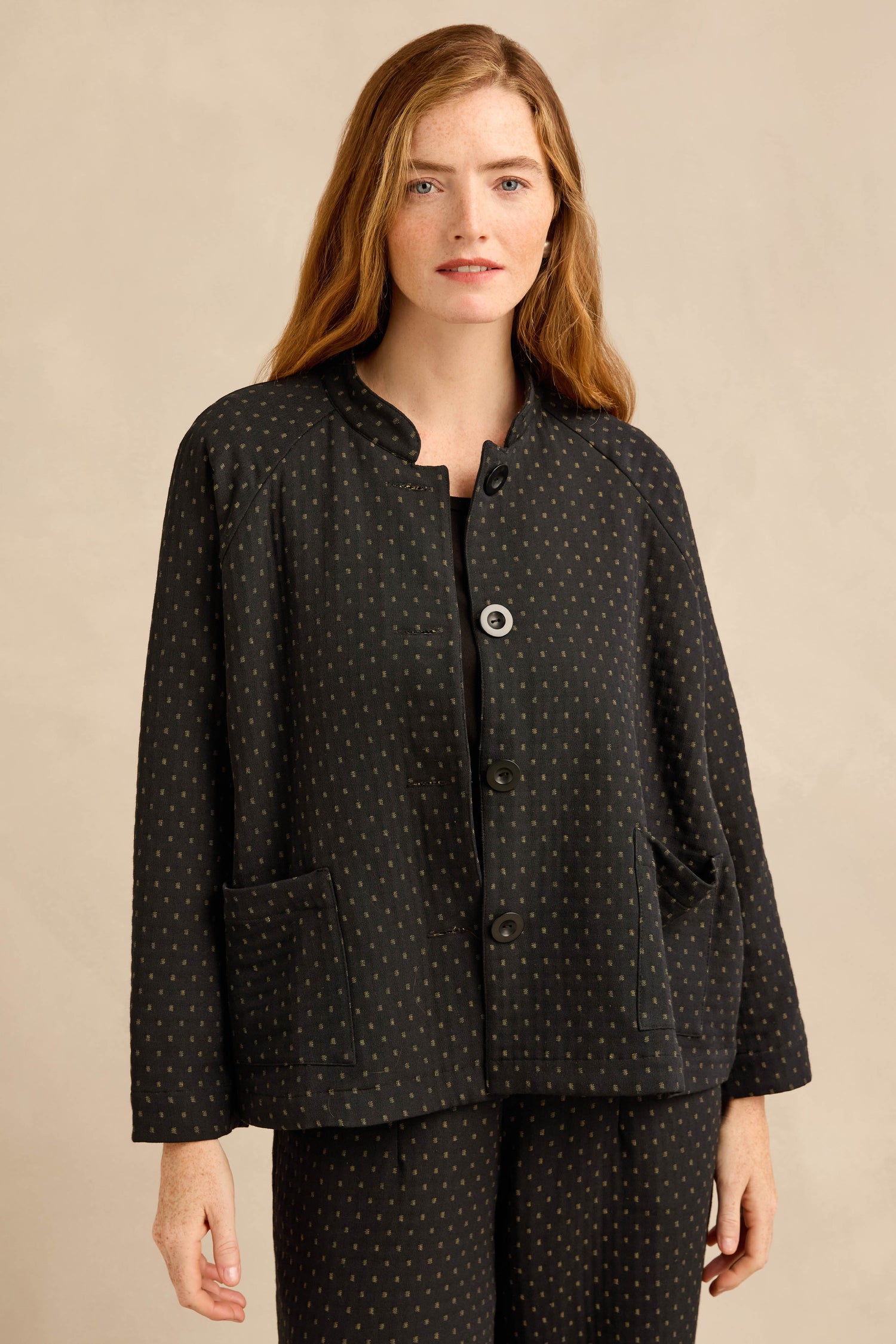 A woman with long, light brown hair wears the Square Jacquard Jacket—workwear-inspired with small dotted patterns and large front pockets—paired with matching pants, standing against a neutral background.