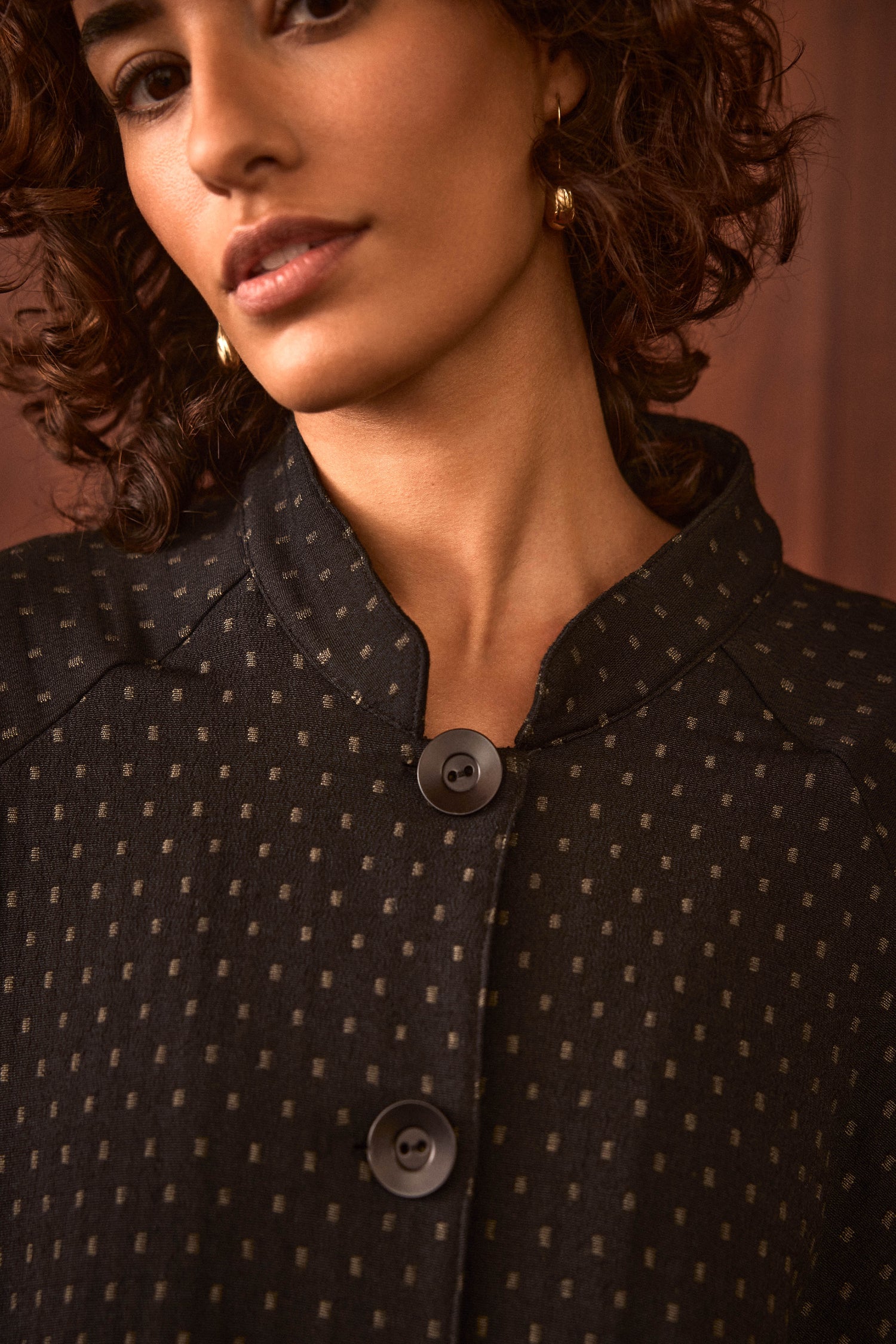 A woman stands against a brown background wearing the Square Jacquard Jacket—a black, cotton-rich jacket with a subtle square pattern and two large buttons.