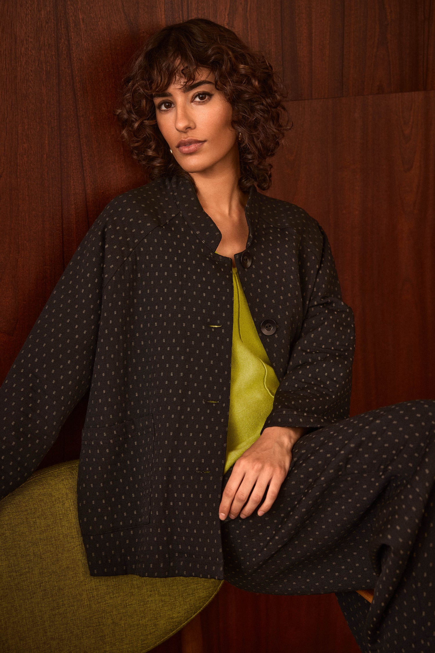 A person with short curly hair sits against a dark wooden wall, wearing the Square Jacquard Jacket—a dark-patterned, cotton-rich piece—layered over a bright green top.