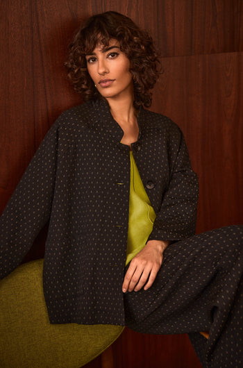 A person with short curly hair sits against a dark wooden wall, wearing the Square Jacquard Jacket—a dark-patterned, cotton-rich piece—layered over a bright green top.