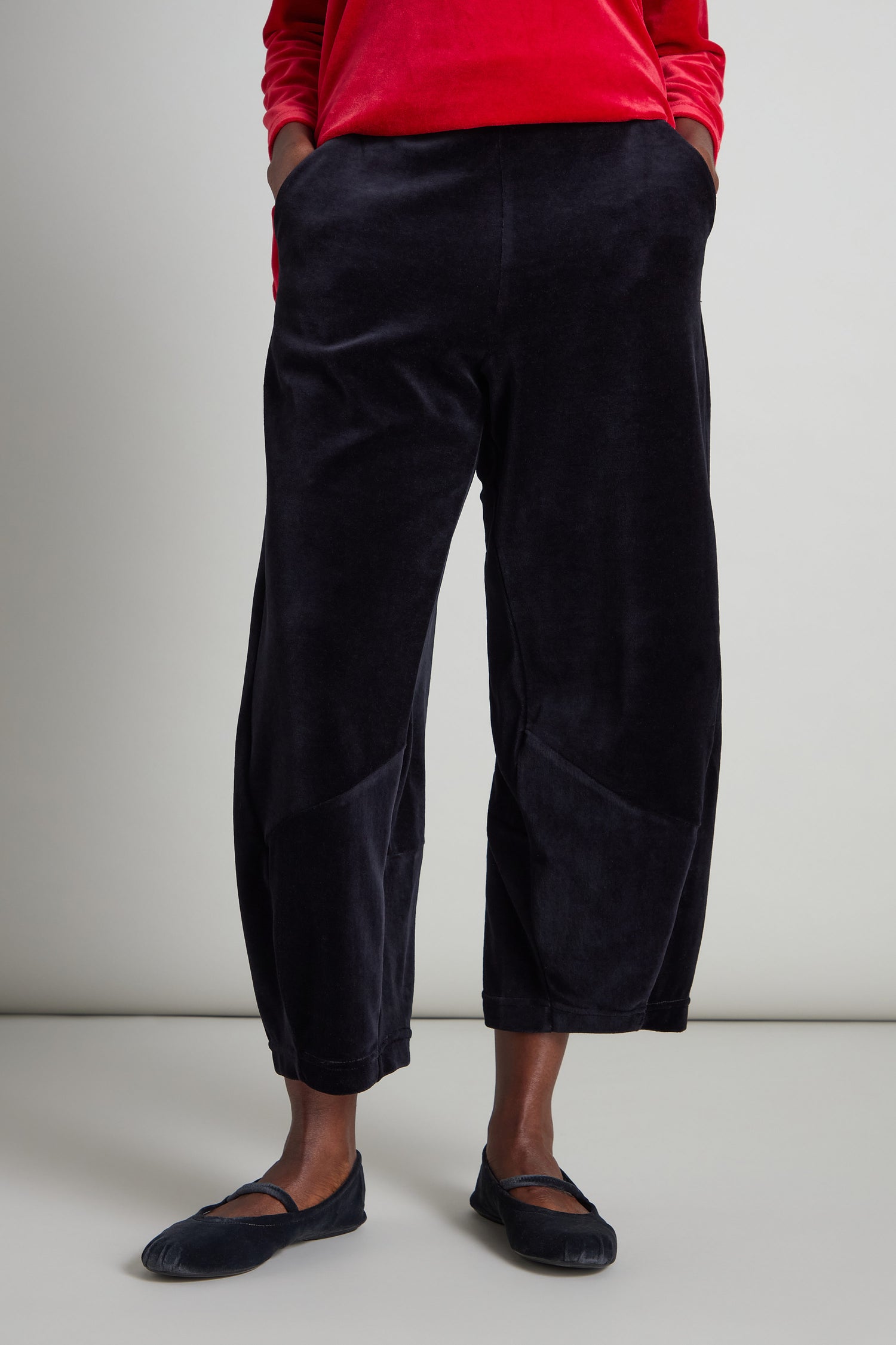 A person in a red long-sleeve shirt and black ballet flats, with hands in pockets, completes their look with the stylish and comfortable Velvet Jersey Bubble Trousers featuring an elasticated waist.