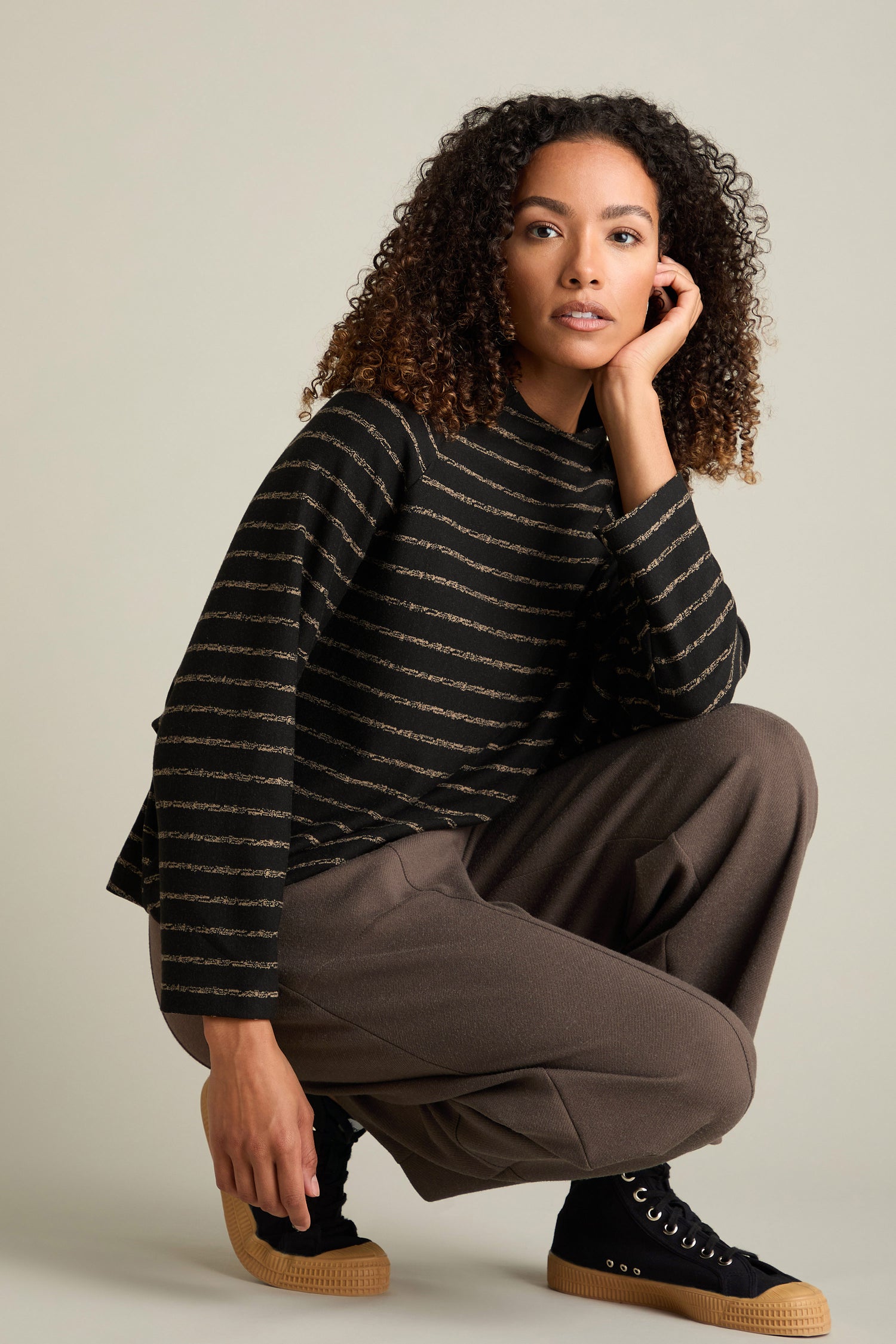 A person wearing a black and striped sweater paired with Marl Jersey Bubble Trousers in brown is squatting against a neutral background, embodying contemporary style.