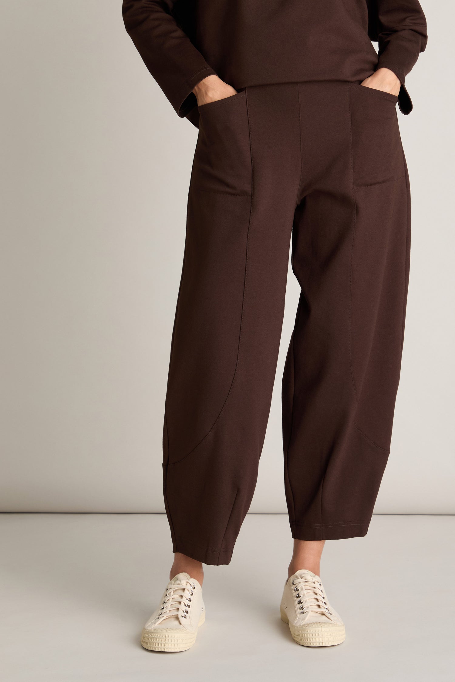 Person wearing flattering loose Ponte Bubble Trouser in brown with an elasticated back and cream-colored sneakers stands against a plain background with their hands in their pockets.