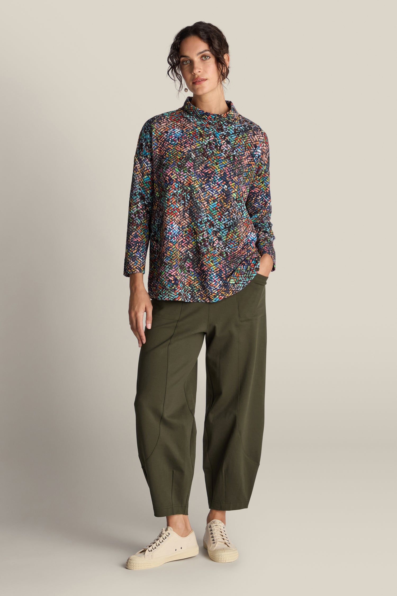 A woman stands against a plain background wearing a multicolored patterned long-sleeve top, olive green Ponte Bubble Trousers, and beige sneakers. One hand is in her pocket, highlighting this stylish year-round staple, the Ponte Bubble Trouser.