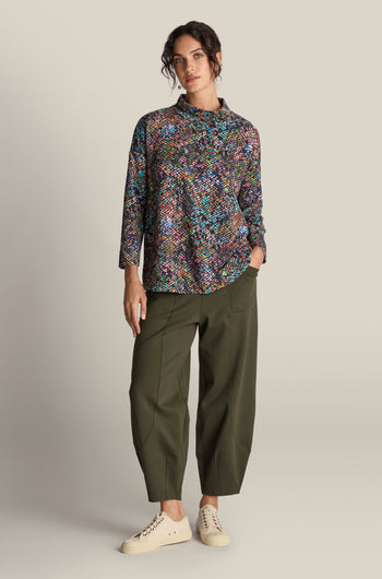 A woman stands against a plain background wearing a multicolored patterned long-sleeve top, olive green Ponte Bubble Trousers, and beige sneakers. One hand is in her pocket, highlighting this stylish year-round staple, the Ponte Bubble Trouser.