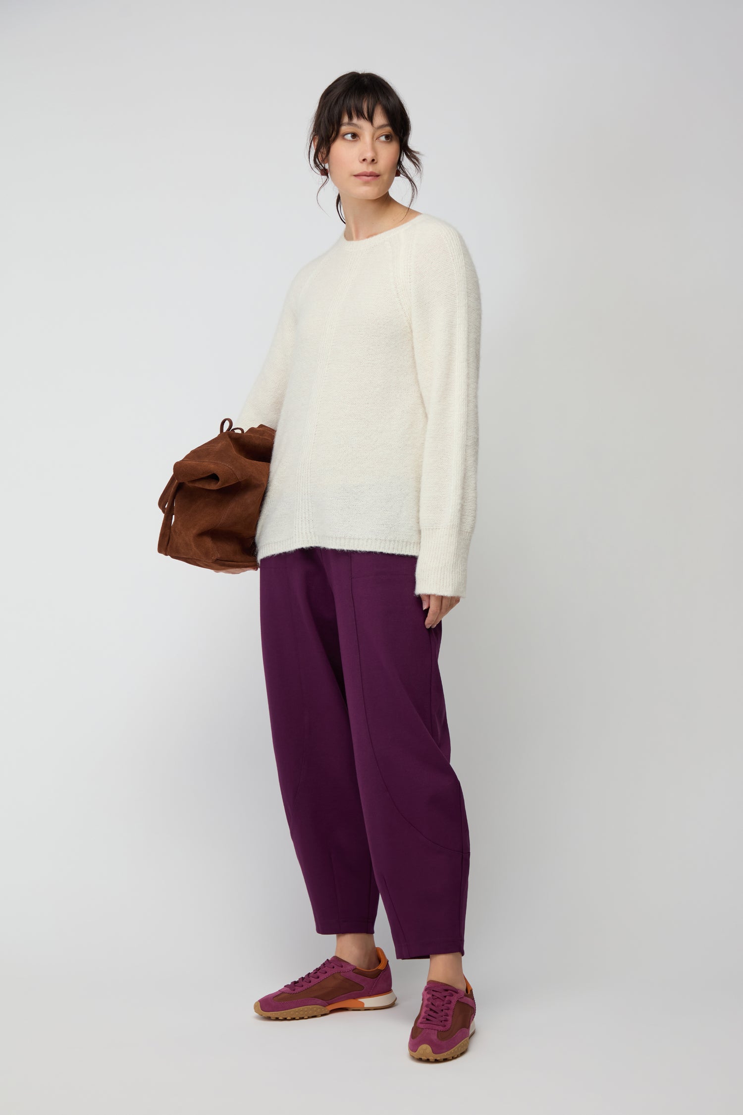 A person stands against a plain backdrop wearing a white sweater, purple Ponte Bubble Trousers with an elasticated waistband, and purple sneakers, holding a brown bag under one arm.