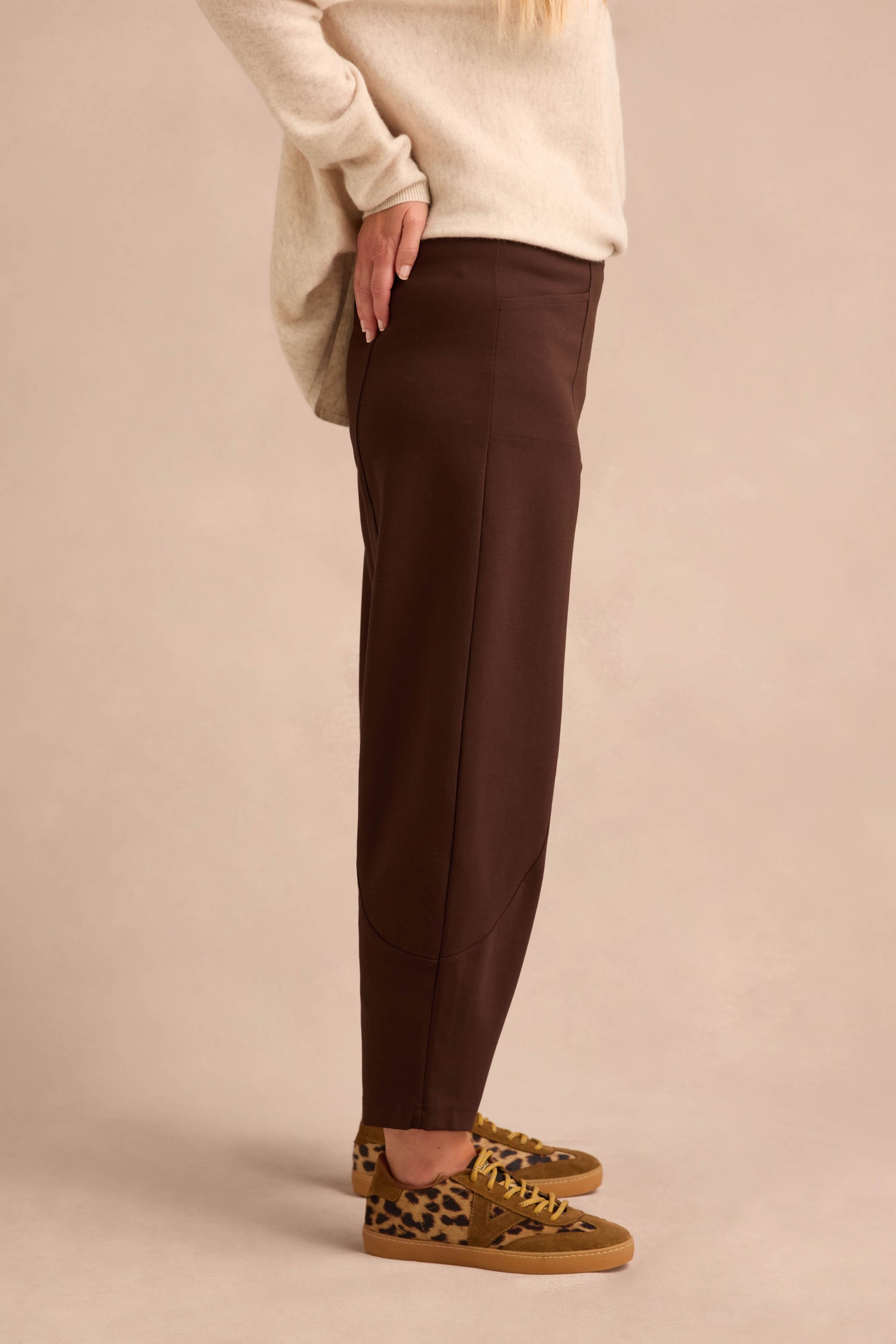 A person stands sideways against a plain beige background wearing Chocolate Ponte Bubble Trousers with a rounded leg, paired with a cream sweater and leopard-print sneakers.