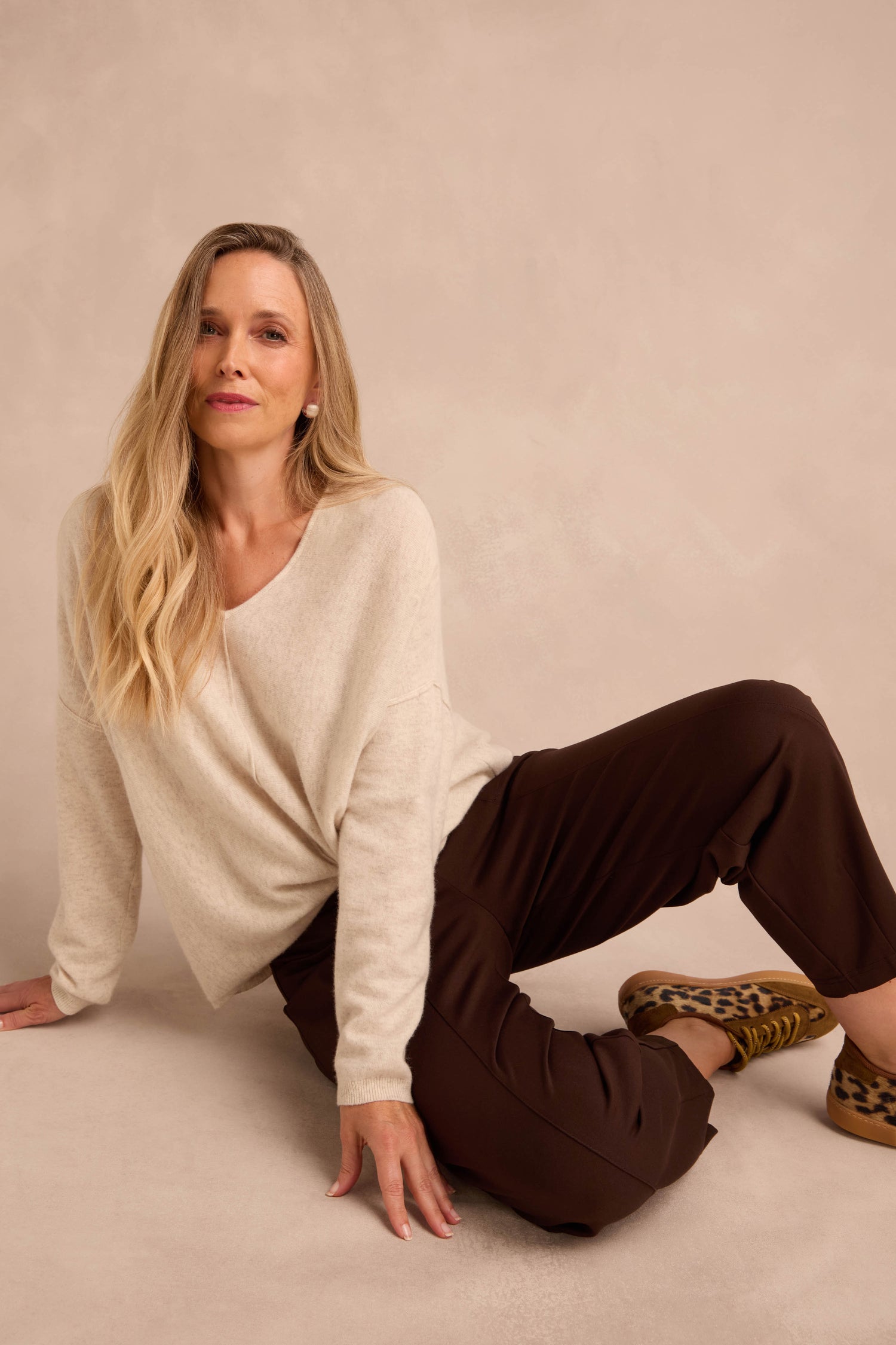 Woman with long blonde hair in a cream sweater and Chocolate Ponte Bubble Trousers sits on the floor against a beige background, wearing leopard print shoes.