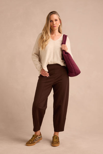 Woman stands against a beige background, wearing a cream sweater, Chocolate Ponte Bubble Trousers, leopard sneakers, and carrying a maroon bag over her shoulder.