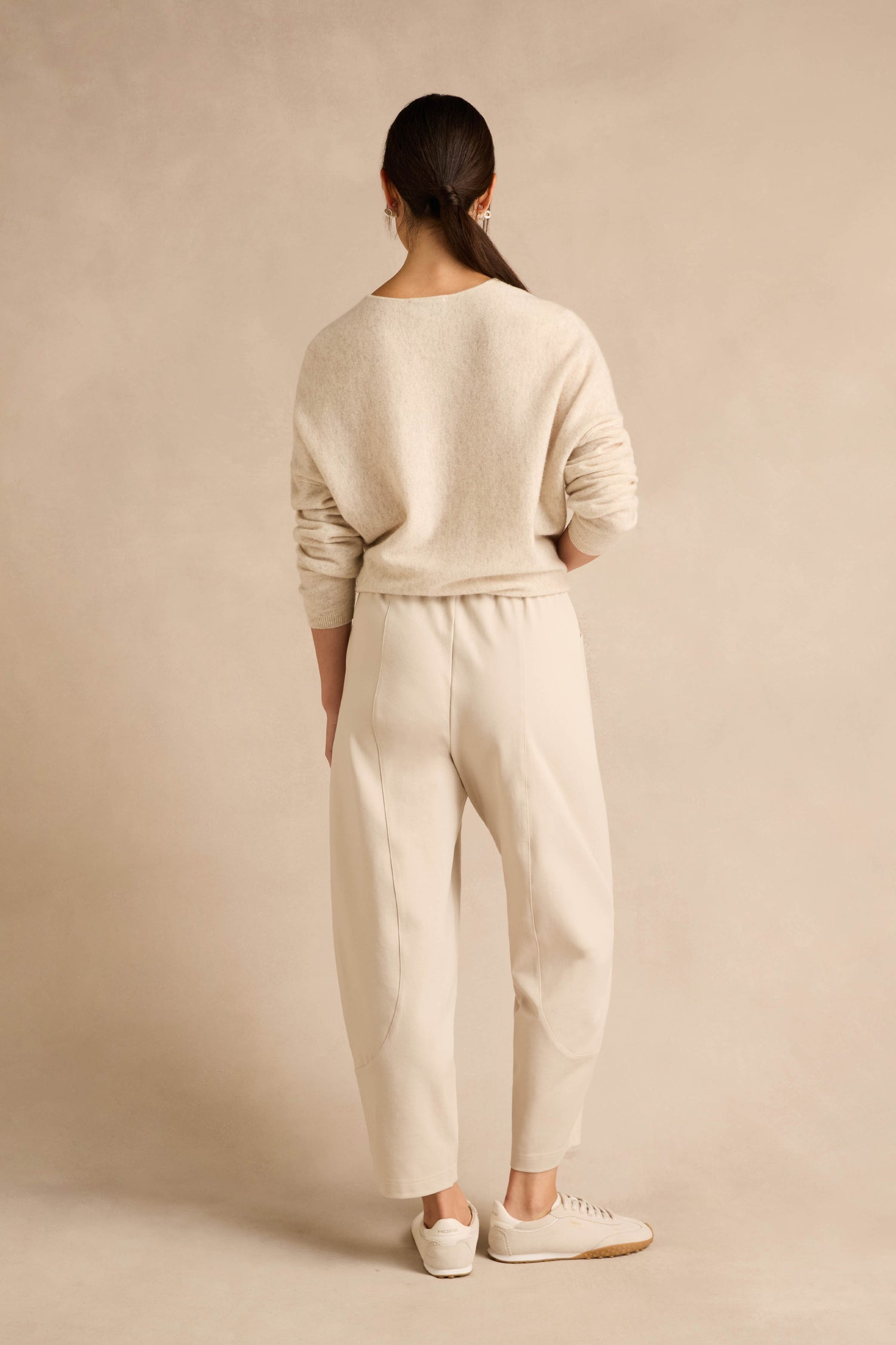 Person standing with back to camera, wearing a light beige sweater paired with Ecru Ponte Bubble Trousers and white sneakers against a neutral background.
