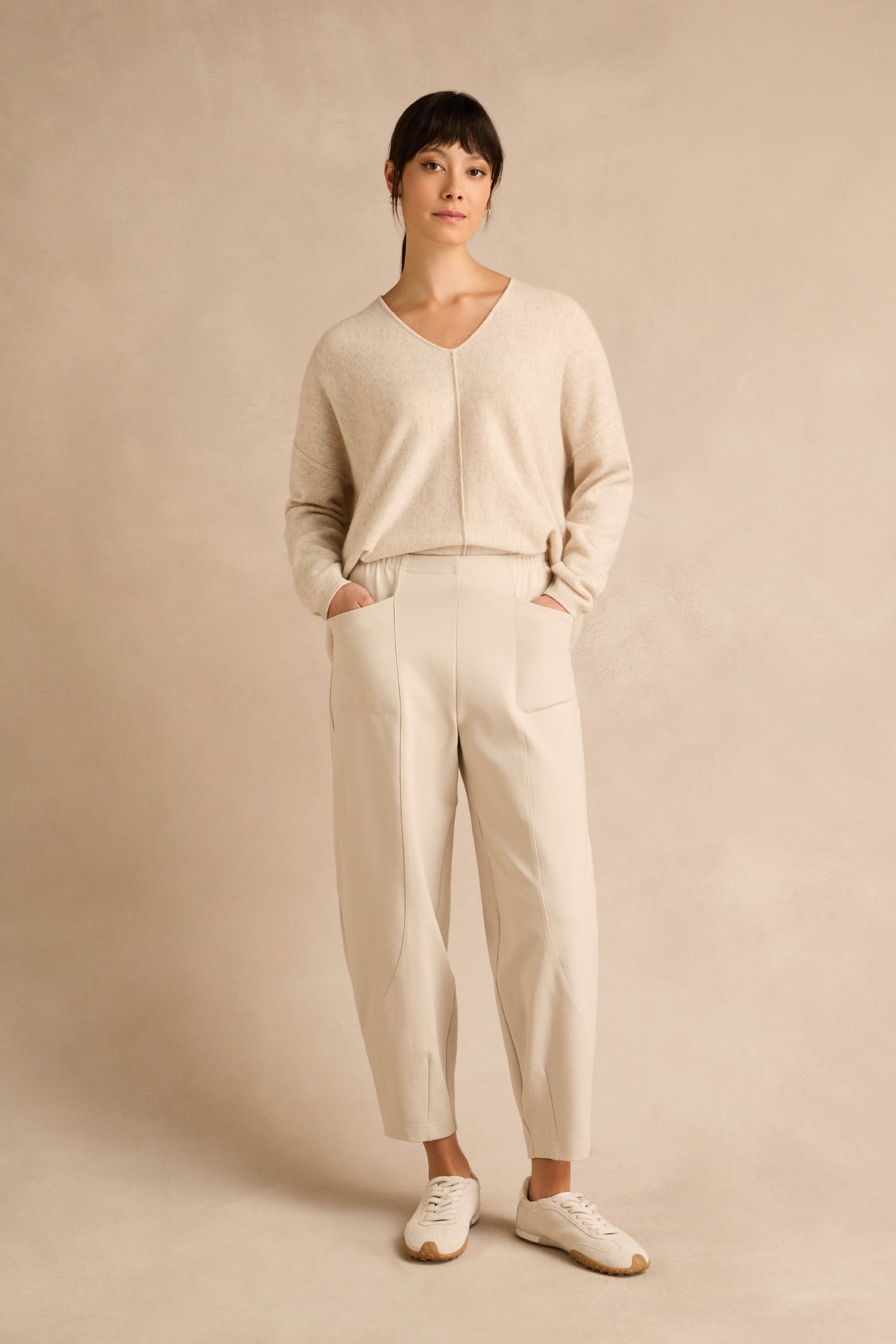 A woman stands against a beige background, wearing a light beige sweater, Ecru Ponte Bubble Trousers in a cream-colored hue that create a subtle bubble silhouette, and white sneakers, with her hands in her pockets.