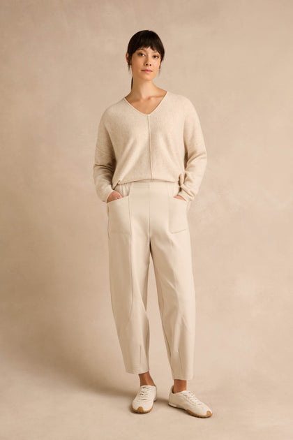 A woman stands against a beige background, wearing a light beige sweater, Ecru Ponte Bubble Trousers in a cream-colored hue that create a subtle bubble silhouette, and white sneakers, with her hands in her pockets.