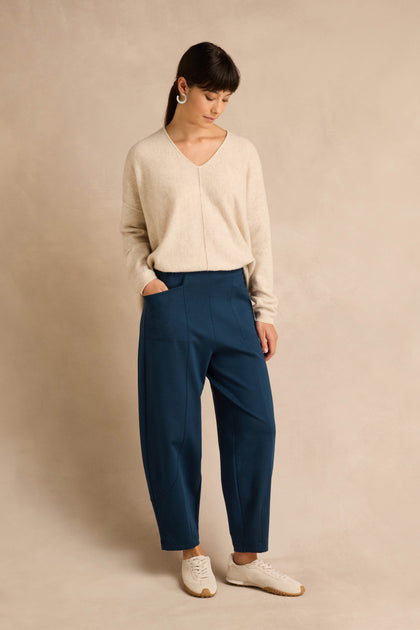 A person stands against a neutral background, wearing a beige sweater, white sneakers, and large earrings, with hands in pockets and head slightly tilted down. They are dressed in the Marine Ponte Bubble Trousers—blue high-waisted trousers with a rounded leg silhouette crafted from soft ponte jersey.