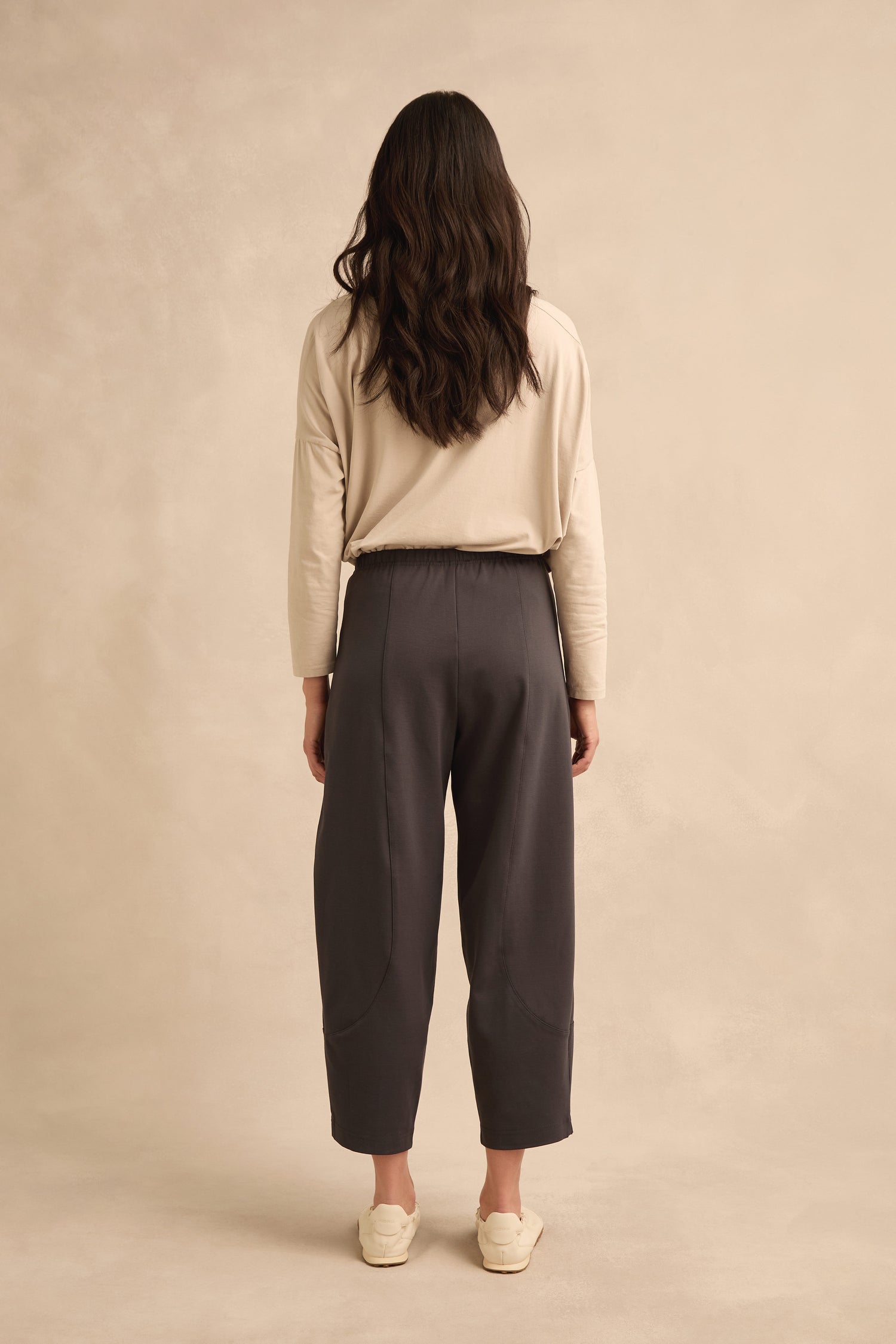 A person with long hair stands facing away, wearing a beige long-sleeve shirt, Slate Ponte Bubble Trousers with an elasticated waist, and cream shoes, against a plain beige background.