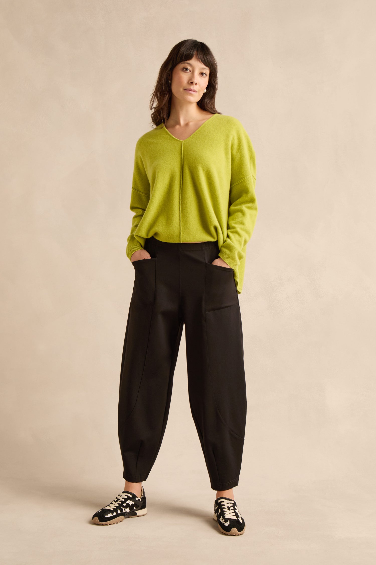 A woman stands with her hands in her pockets, wearing a lime green sweater, Black Ponte Bubble Trousers, and black-and-white sneakers against a beige background.