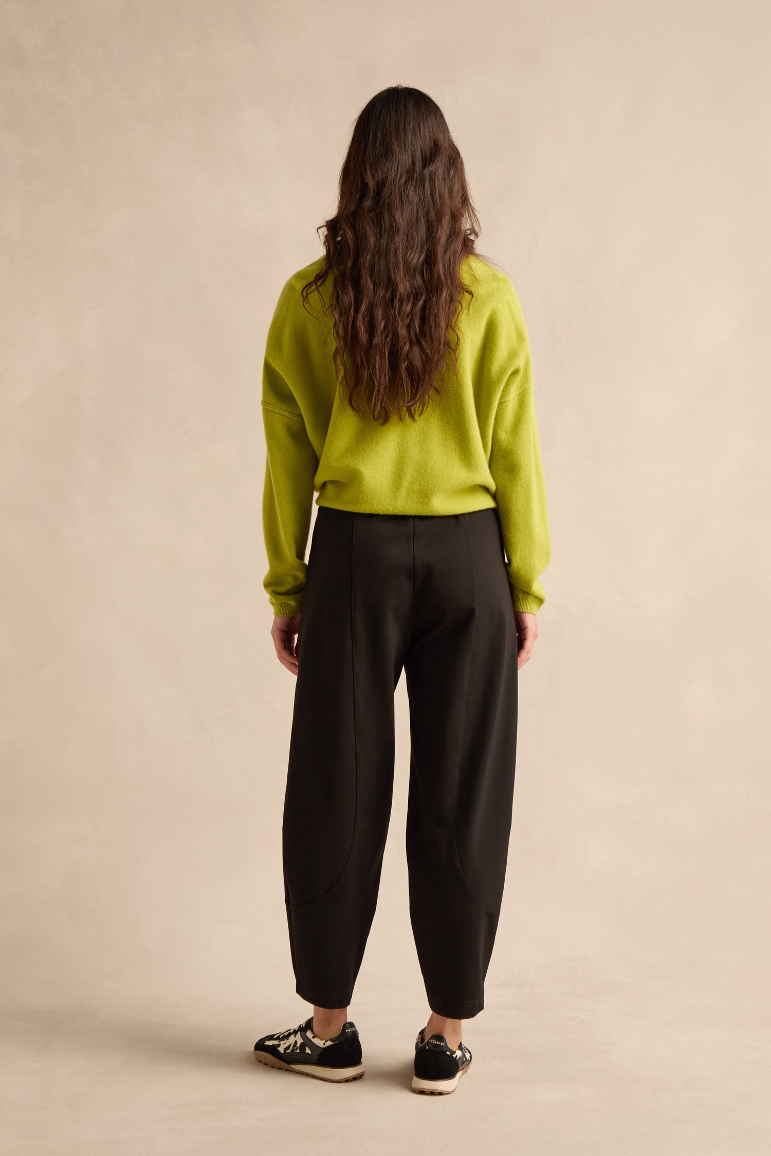 A person stands facing away from the camera, wearing a lime green sweater, Black Ponte Bubble Trousers, and black sneakers against a neutral beige background.