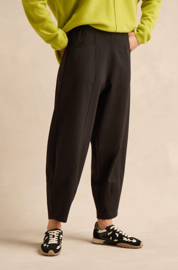 Wearing the Black Ponte Bubble Trousers, paired with a lime green sweater and black-and-white sneakers, the model stands against a plain beige background. The photo focuses on the lower half of the body, emphasizing the trousers’ contemporary bubble silhouette.