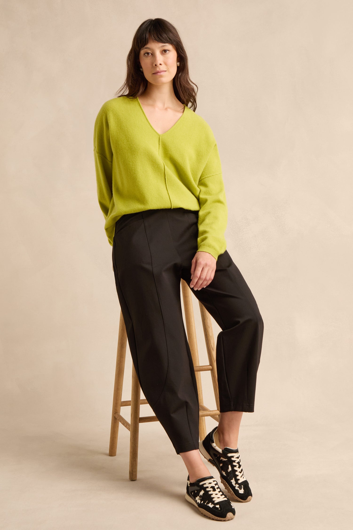 Woman wearing a lime green sweater, Black Ponte Bubble Trousers, and black sneakers sits on a wooden stool against a neutral background.