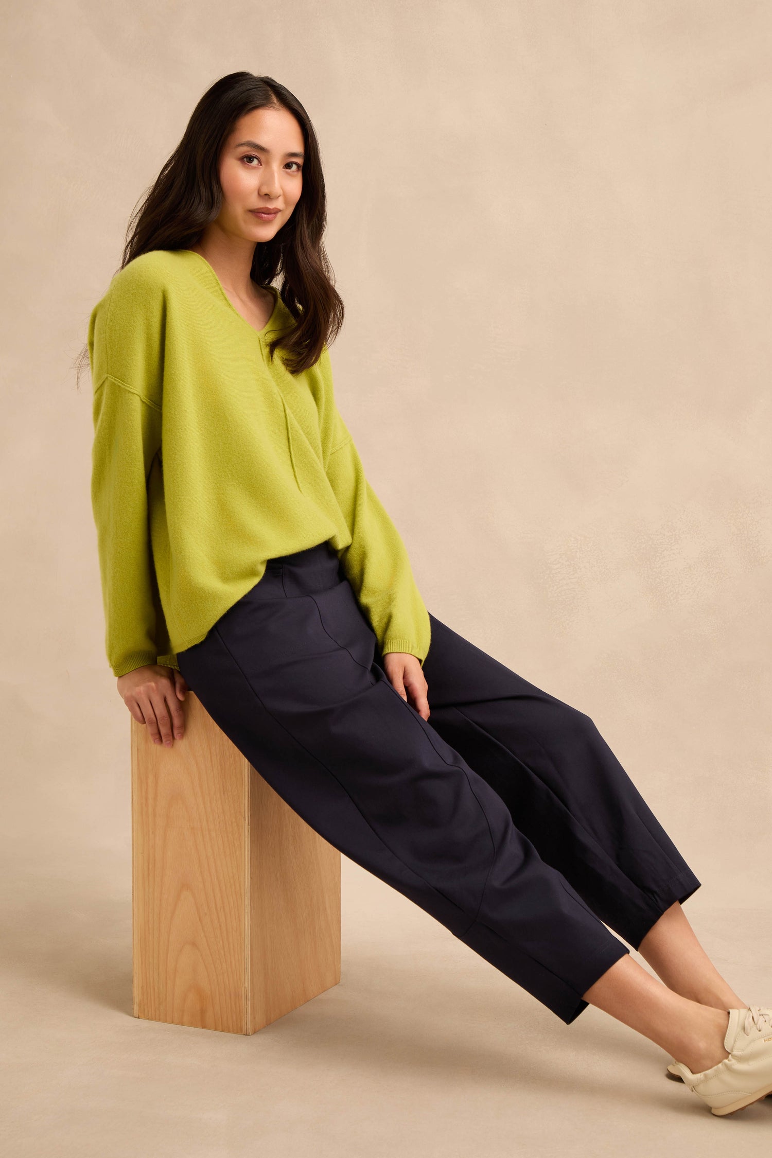 A woman with long dark hair sits on a wooden block, wearing Midnight Ponte Bubble Trousers with a lime green sweater and beige shoes against a neutral background.
