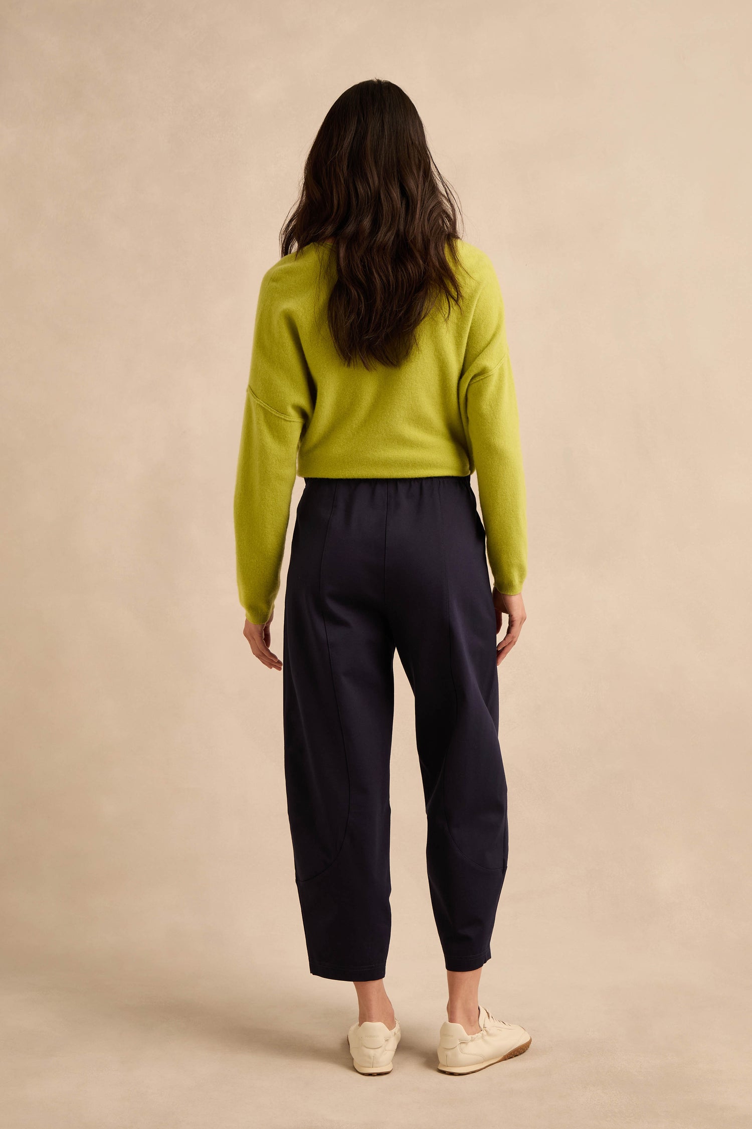 Person standing facing away from the camera, wearing a lime green sweater, Midnight Ponte Bubble Trousers with a rounded leg silhouette in navy, and white sneakers, against a beige background.