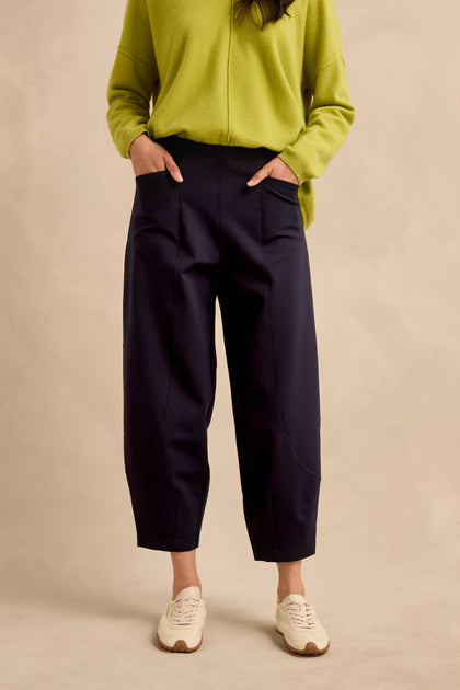 A person wearing a lime green long-sleeve top and the Midnight Ponte Bubble Trousers—loose black pants featuring large front pockets and a rounded leg silhouette—stands with hands in their pockets, paired with white sneakers against a plain beige background.