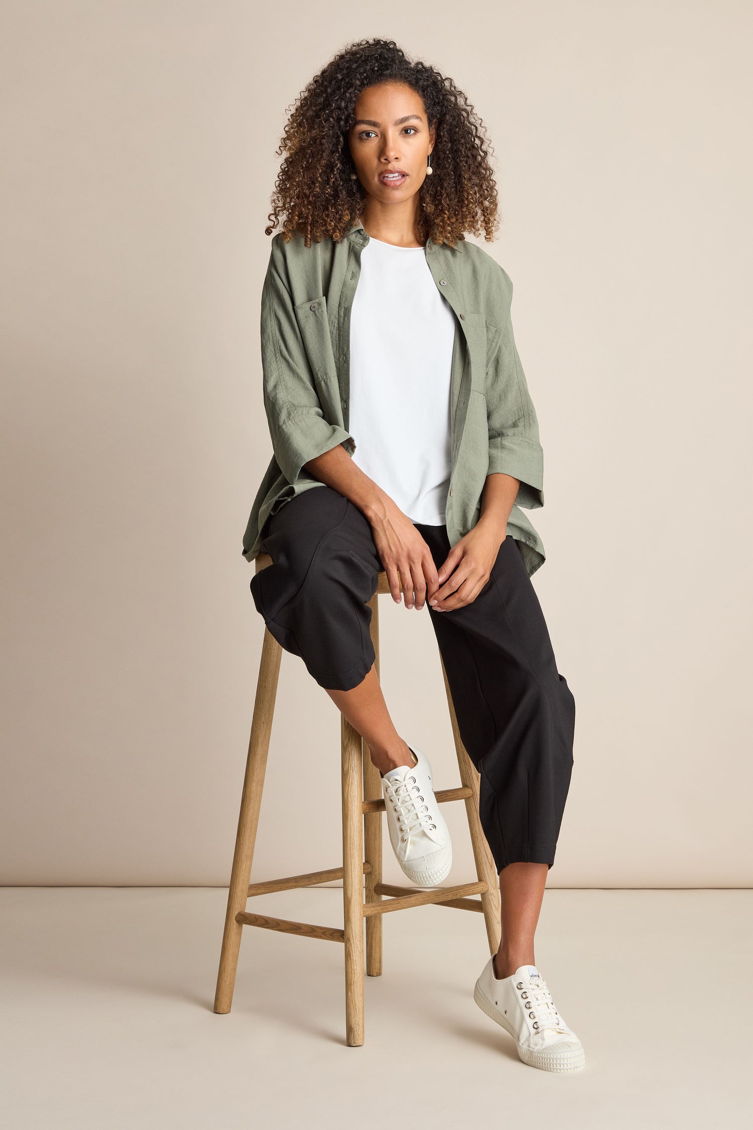 A woman with curly hair sits on a wooden stool. She wears a green jacket, white shirt, and the Ponte Bubble Trouser, which features an elasticated waistband. Her ensemble is completed with white sneakers. The background is plain and beige.