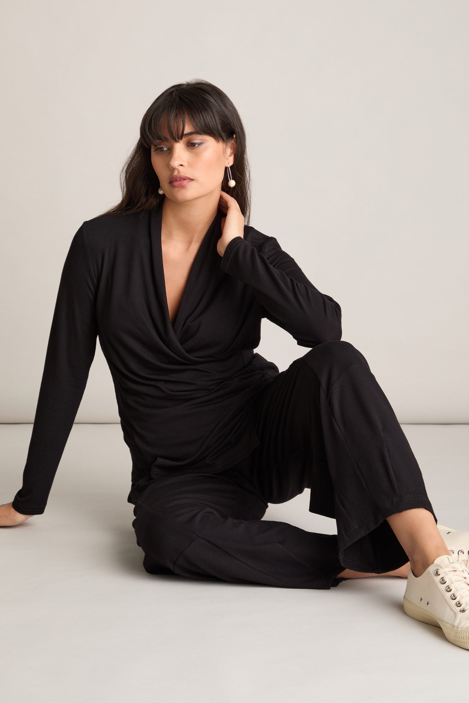 A person sits on the floor with one leg folded, showing off a chic co-ord look that includes a black long-sleeve top and Fluid Crepe Bubble Trousers featuring an elasticated waist. White sneakers complete her outfit as she looks to the side, touching her neck with one hand.