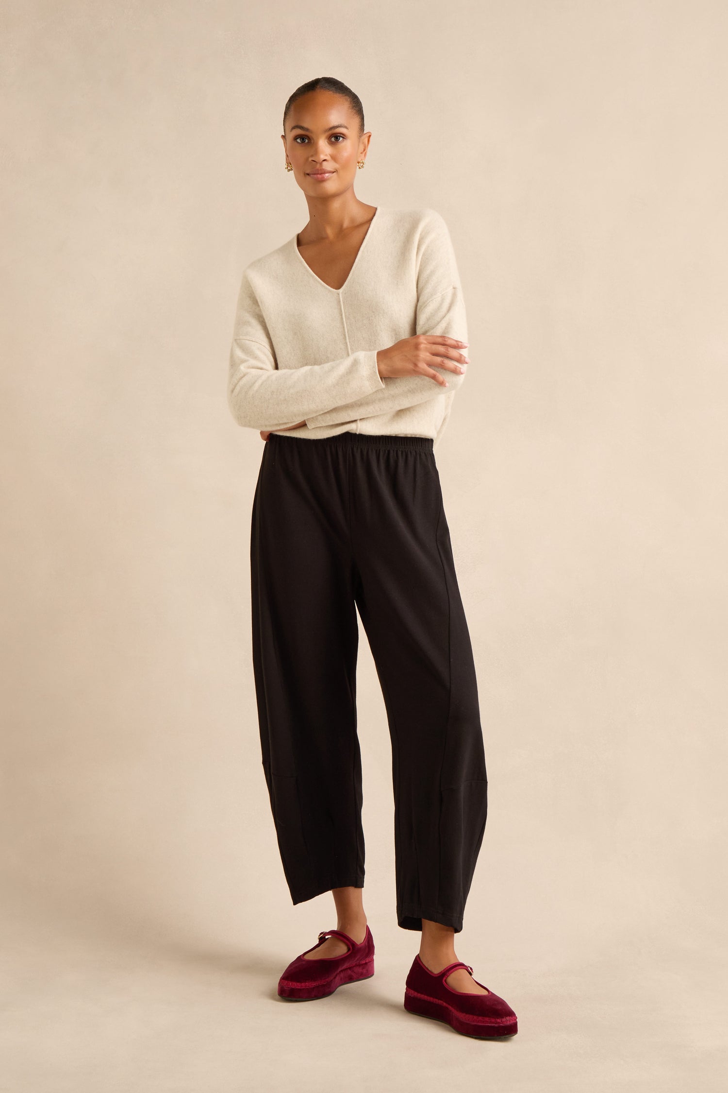 A person stands facing forward, wearing a beige V-neck sweater, Fluid Crepe Bubble Trouser with a rounded leg and slim bubble silhouette, paired with burgundy velvet shoes against a neutral background.